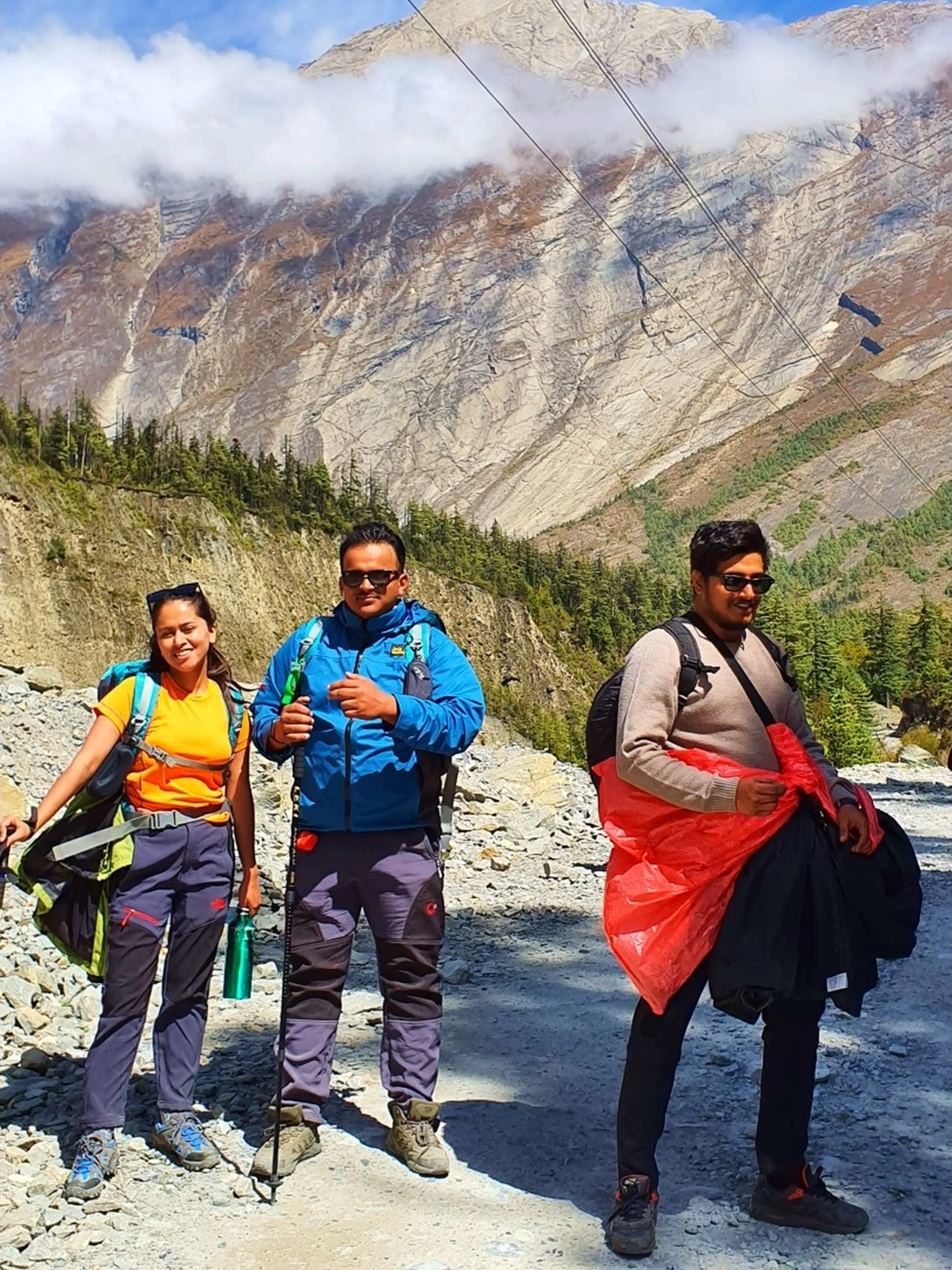 Three Nepali trekkers stand together on a wide rocky mountain trail in Manang, Nepal with a magnificent cloud-draped Himalayan rock face as backdrop - a young woman in an orange tee, green jacket, and trekking trousers smiling on the left - a man in a blue trekking jacket and dark trousers in the center - another in a beige sweater with sunglasses and red plastic bags on the right - the enormous grey, rust-brown, and white-streaked cliff mountain fills the background, pine forest visible below the cloud line - brilliant blue sky with wispy cirrus clouds above - a vibrant and confident Nepal Annapurna Circuit trekking trail group photograph in the high-altitude Manang valley, showcasing the raw and imposing mountain scale of trekking between Chame and Pisang