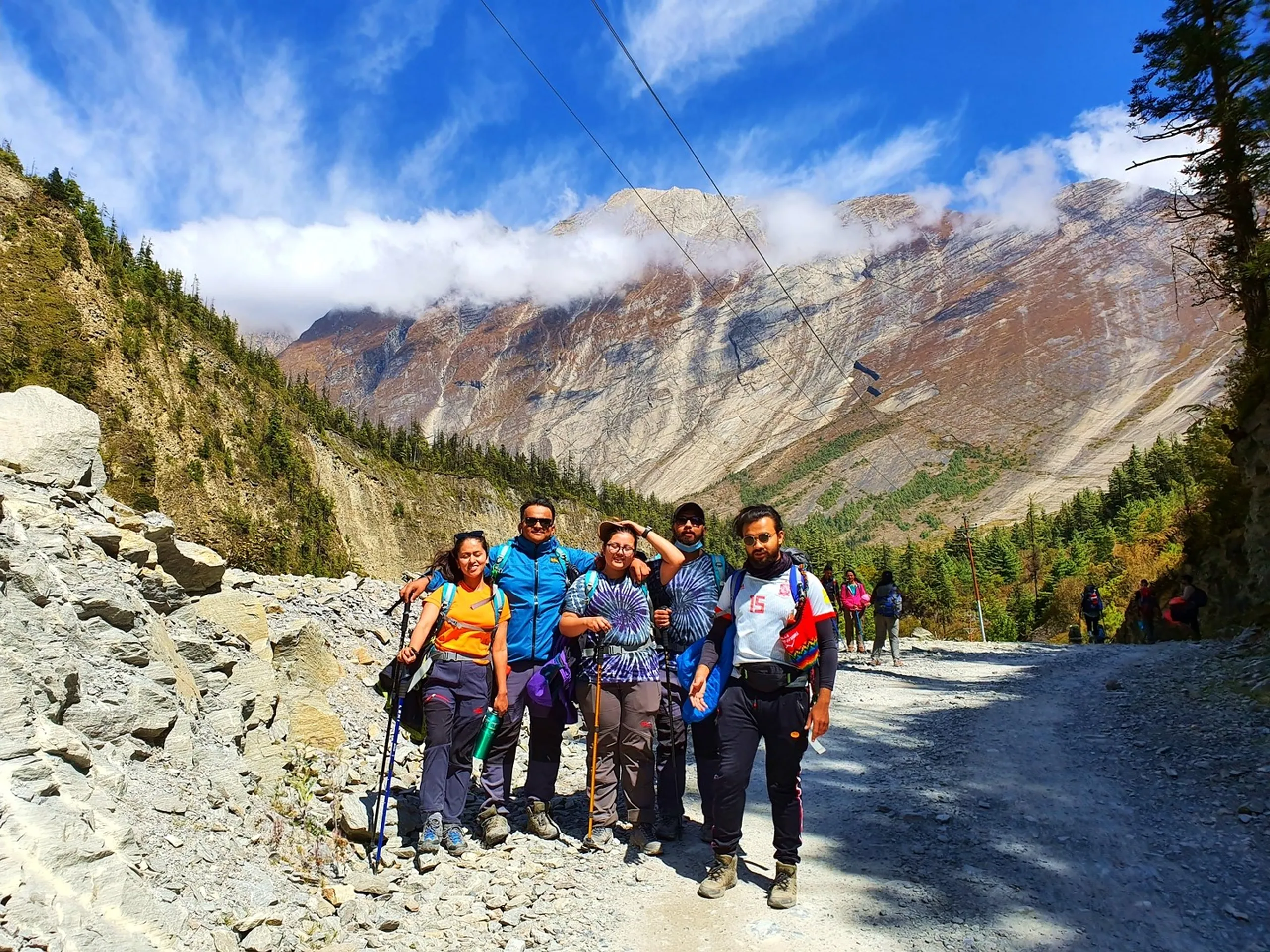 Five young Nepali trekkers stand together on a rocky mountain trail on the Annapurna Circuit in Manang, Nepal - wearing colorful trekking gear including a blue jacket, purple tie-dye shirt, Adidas number 15 football jersey, orange top and green jacket combination - some holding trekking poles - laughing, smiling, and gesturing playfully - a dramatic wall of towering grey and rust Himalayan cliff rock draped in low-hanging white cloud fills the entire background - pine forest visible on the valley floor - a brilliant blue sky with wispy cloud above - other trekkers visible further back on the trail - a joyful and vibrant Nepal trekking group trail photo capturing mountain friendship and adventure on the Annapurna Circuit, on the way to Ice Lake Manang
