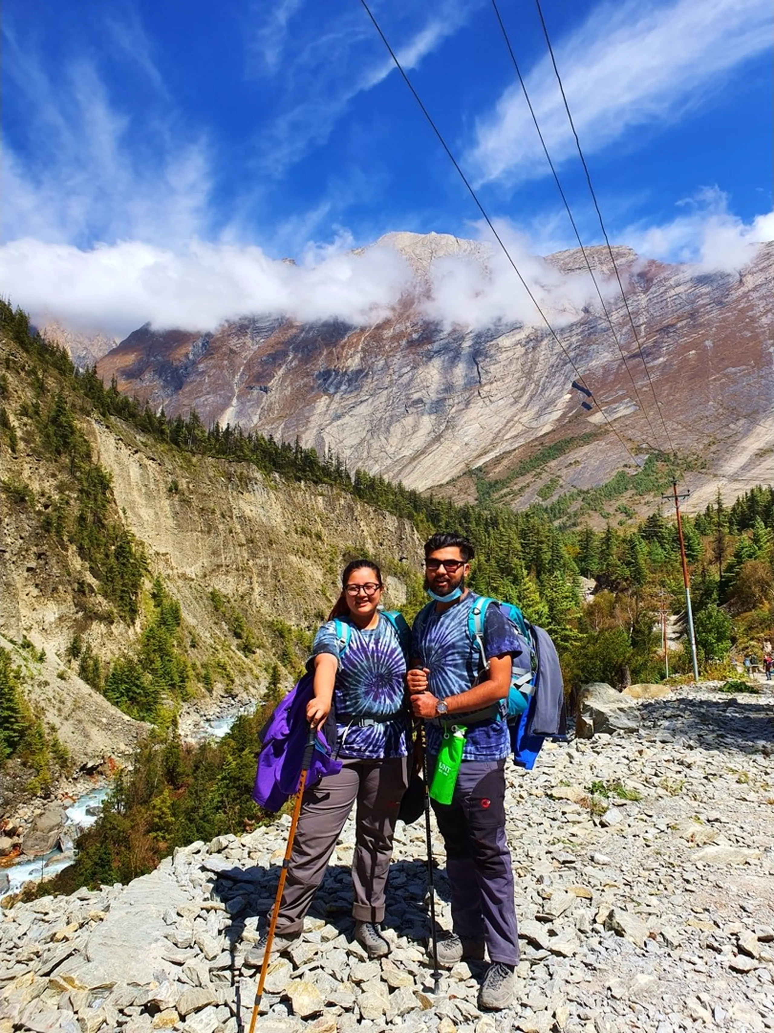Two Nepali trekkers wearing matching purple and grey tie-dye t-shirts stand side by side on a rocky mountain trail in Manang, Nepal, smiling warmly - the woman on the left wearing glasses, a purple jacket, grey trousers, and blue trail shoes, holding a wooden trekking pole - the man on the right wearing sunglasses, a blue-grey jacket, dark trousers, and a green water bottle holster - both carrying loaded backpacks - a dramatic broad Himalayan valley scene behind them with a pale grey and rust-toned mountain ridge wreathed in low white cloud - a turquoise glacial river rushing through the gorge below visible to the left - pine forest and autumn-colored trees on the valley slopes - a beautiful and intimate Nepal couple trekking portrait on the Annapurna Circuit trail to Manang and Ice Lake
