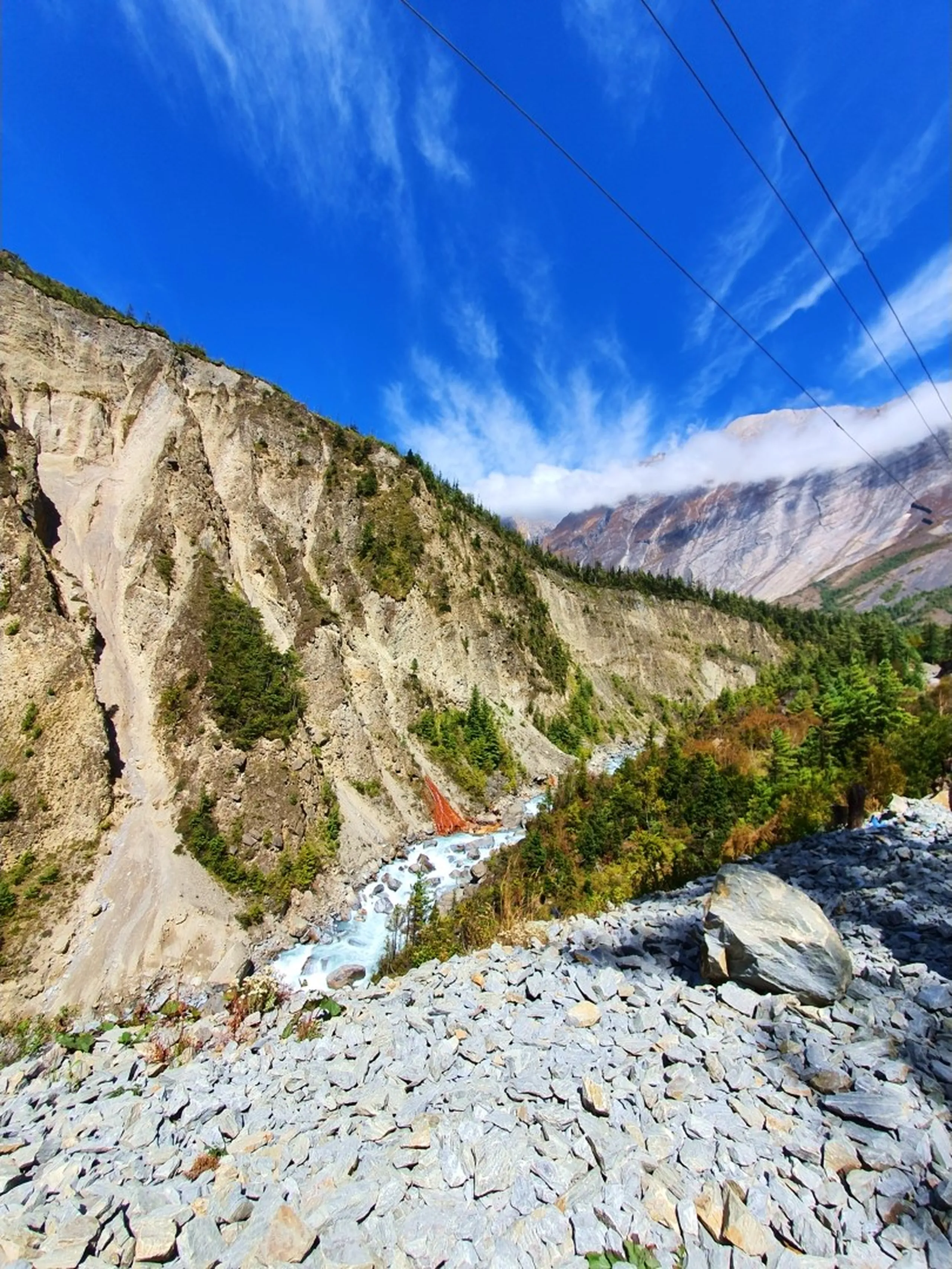 A sweeping wide-angle landscape photograph of the Marsyangdi River valley on the Annapurna Circuit trek route to Manang, Nepal - a turquoise-white glacier-fed river rushes through a broad rocky valley floor strewn with grey shale and boulders - an unusual vivid orange-red mineral spring or geological formation stains the cliffside and riverbank on the left - steep eroded sandy and rocky valley walls rise on both sides, dotted with pine and fir trees - utility wires cross diagonally across the frame - deep blue sky with feathery cirrus clouds above - a dramatic and geologically unique Nepal Himalayan river valley landscape photograph on the Annapurna Circuit trekking trail, perfect for Nepal nature photography and mountain geology travel blog content