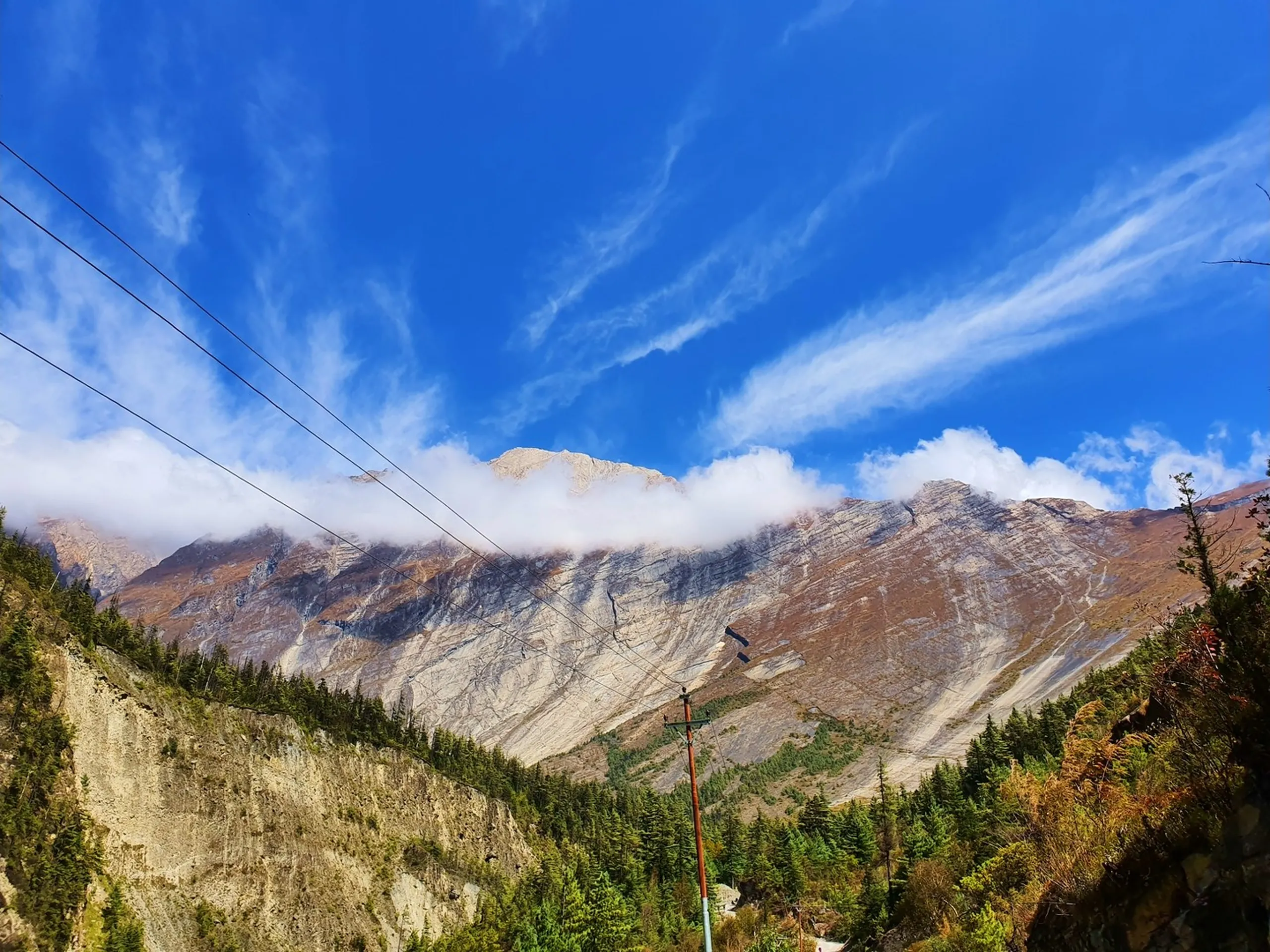 A wide upward-angle landscape photograph of a dramatic Himalayan mountain ridge in Manang, Nepal - a long wall of bare grey and rust-brown rock ridgeline fills the lower frame, laced with scree gullies and pale geological striations - a thick band of low white cloud wraps around the mid-section of the mountain - above the cloud, a rocky summit peak emerges into a vivid deep blue sky laced with wispy cirrus clouds - pine and fir forest cloaks the lower slopes - utility poles and power lines cross the lower frame - a majestic and atmospheric Nepal mountain landscape photograph on the Annapurna Circuit trail in Manang district, showcasing the raw grandeur of the high-altitude Himalayan terrain between Chame and Pisang, ideal for Nepal mountain photography blog content