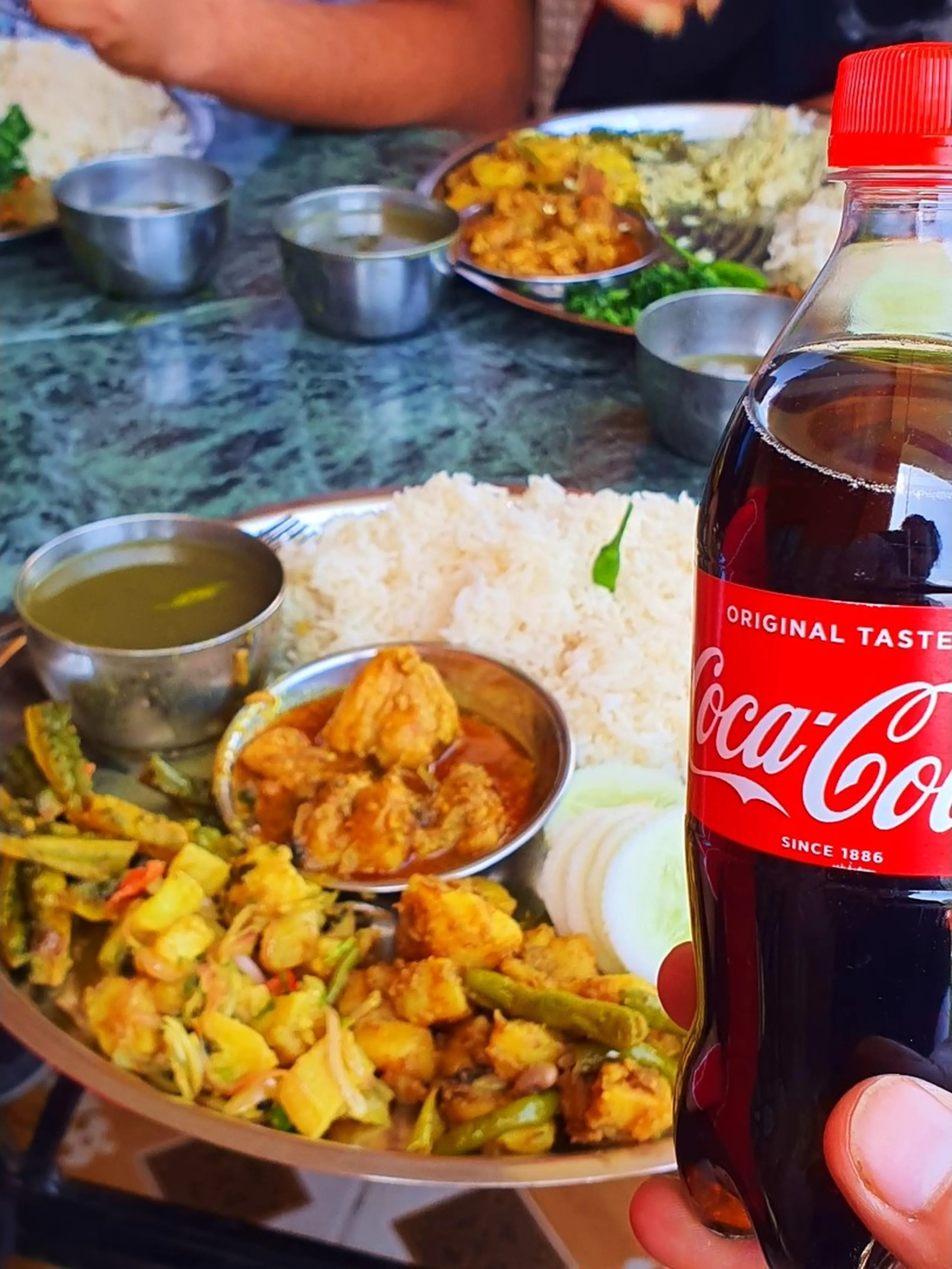 A close-up overhead shot of a traditional Nepali dal bhat thali meal served on a steel plate - featuring steamed white rice, golden vegetable curry with green beans and potatoes, a small bowl of chicken curry, lentil dal in steel bowls, leafy green saag, and sliced onion garnish - with a hand holding a Coca-Cola bottle in the foreground, set on a green marble table with other thali plates visible in the background, photographed during a lunch stop near Kurintar on the way to Manang, Nepal - perfect food photography for Nepal trekking food and dal bhat culture blogs