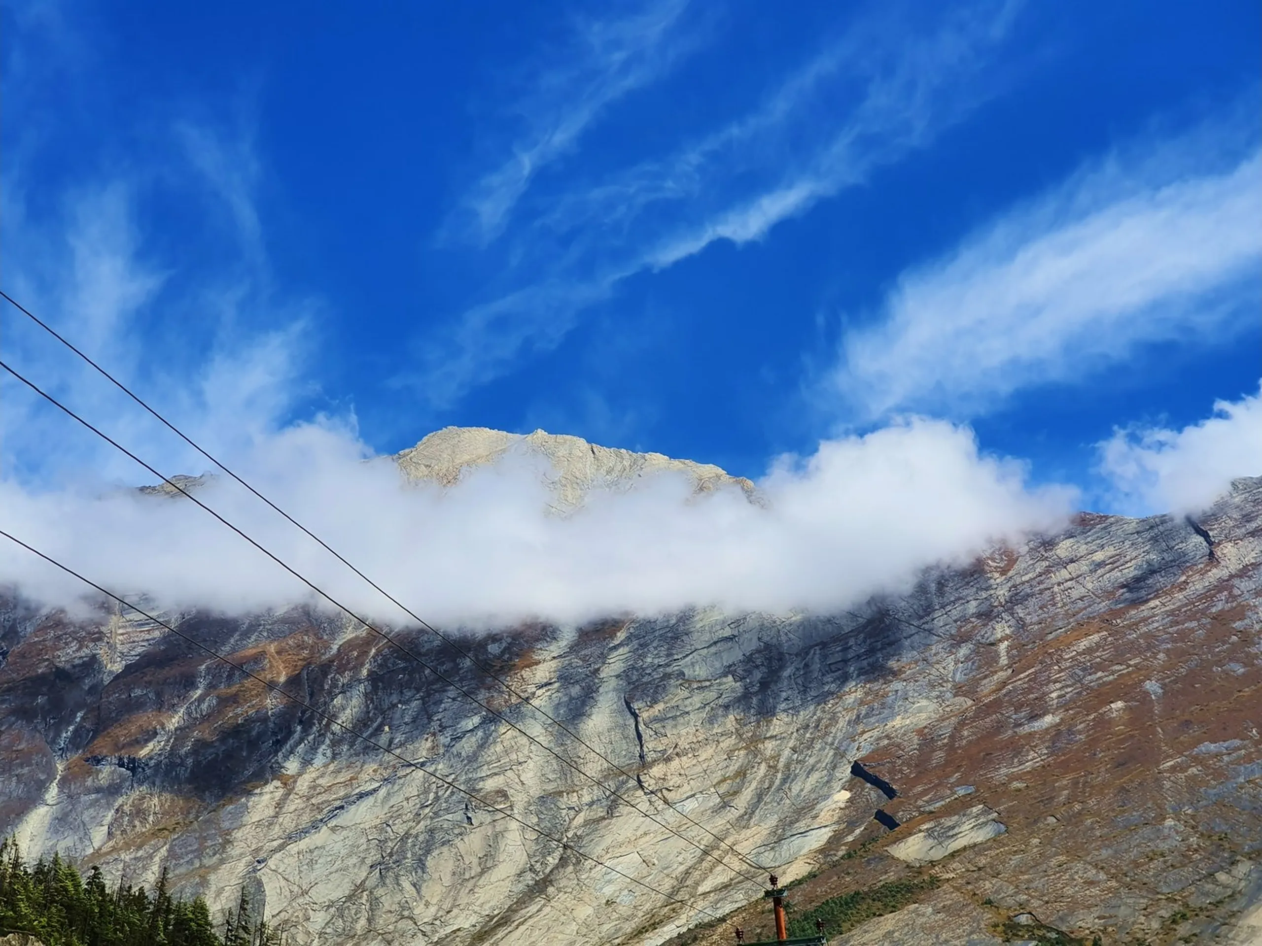 A dramatic upward-angle close-up landscape photograph of a towering Himalayan peak in Manang, Nepal - a massive pale grey and white-streaked rock face scored with geological fault lines and dark erosion channels fills the entire lower frame - a thick horizontal band of brilliant white cloud wraps around the mountain mid-section like a collar - above it, a rugged rocky summit with patches of snow peers into a vivid cobalt blue sky streaked with high cirrus cloud - utility wires cross the lower frame diagonally - pine treetops visible in the bottom right corner - a breathtaking and imposing Nepal Himalayan peak close-up photograph on the Annapurna Circuit trekking trail near Pisang, Manang - perfect for Nepal mountain photography, Annapurna trekking scenery, and Himalayan landscape blog content