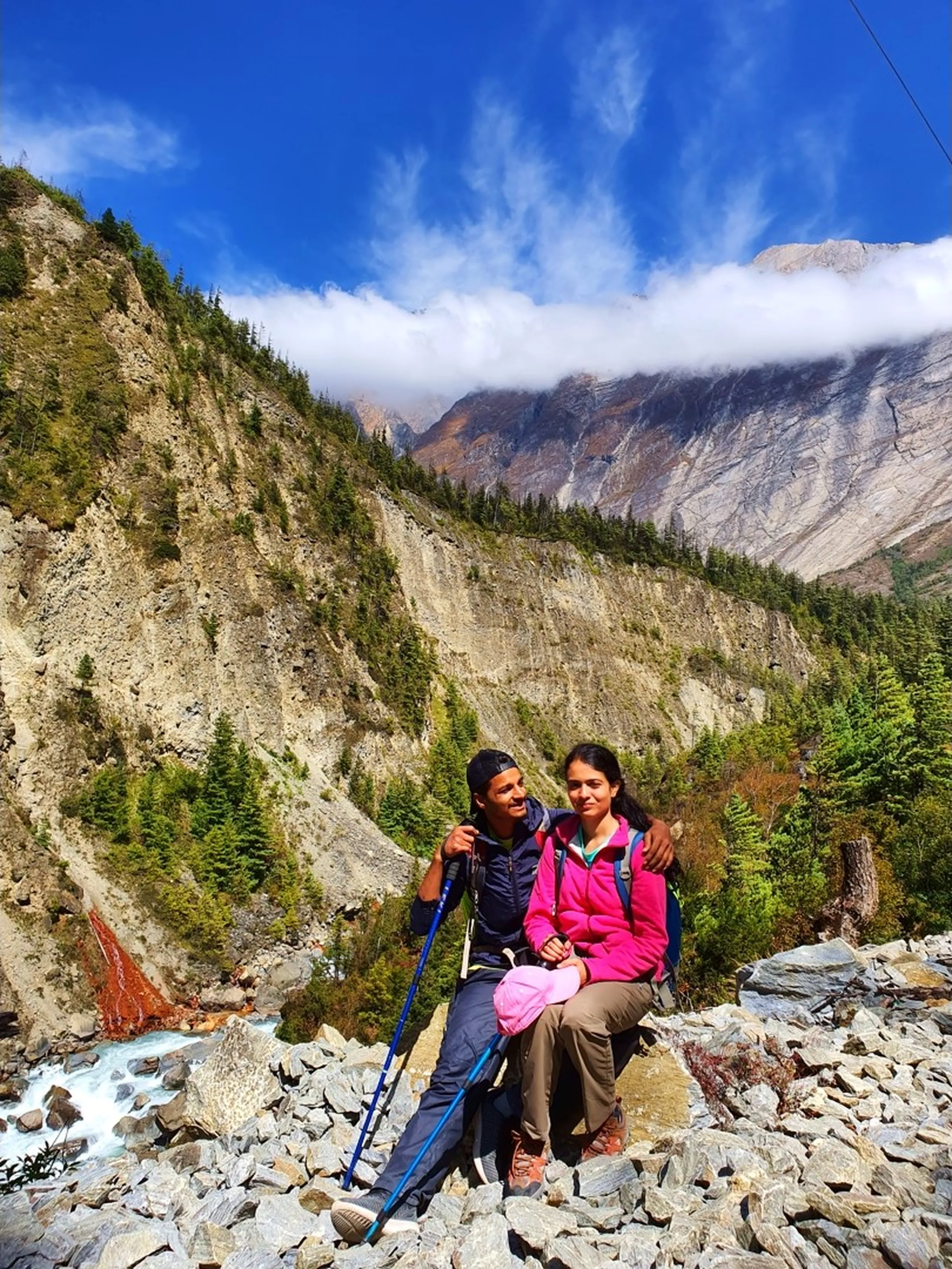 A young Nepali couple sit closely together on a rocky outcrop beside the turquoise Marsyangdi River in Manang, Nepal - the young man in a navy trekking jacket holding a blue trekking pole, the young woman in a bright pink fleece jacket holding a pink hat - both smiling softly and relaxed - dramatic grey and rust-toned Himalayan cliff faces draped in low white cloud filling the background - a vivid orange-red mineral spring staining the cliff on the left - green pine forest clinging to the lower slopes - a beautiful and intimate Nepal couple trekking portrait beside a glacial mountain river on the Annapurna Circuit, capturing the romance and adventure of trekking together in the Himalayas, Manang, Nepal