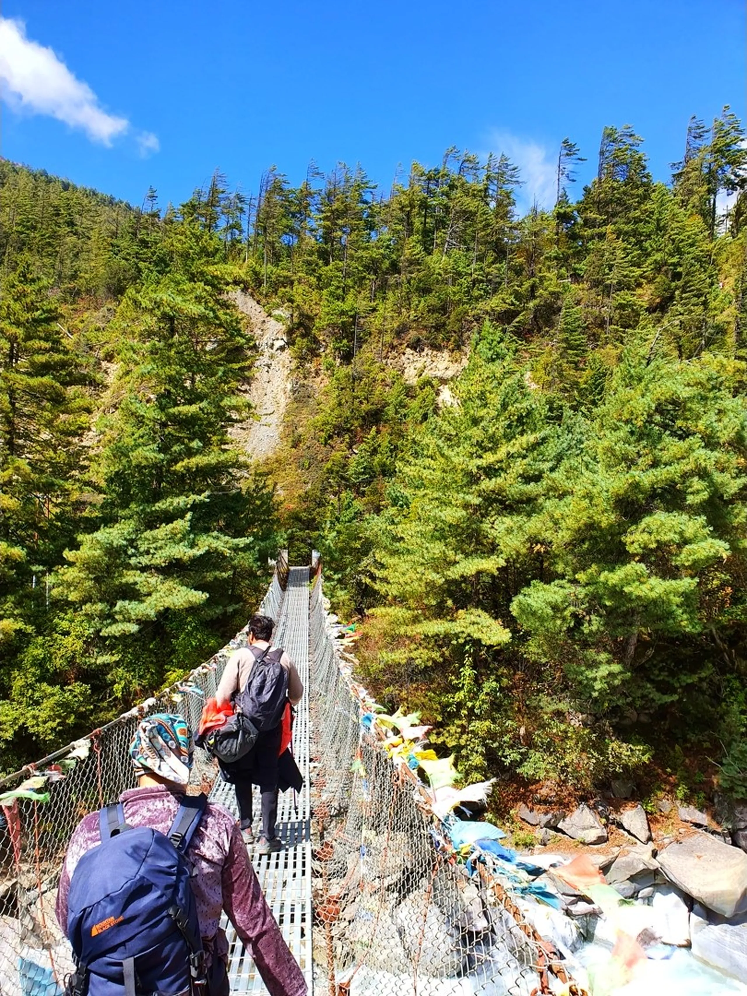 A first-person walking perspective photograph on a steel mesh suspension bridge over a Himalayan river in Manang, Nepal - a male trekker with a backpack and red plastic bags walks ahead towards the far end of the bridge - colorful Tibetan Buddhist prayer flags in red, yellow, blue, green, and white are tied densely along the bridge railings on both sides - dense green pine and fir forest with autumn-tipped foliage fills the background - blue sky with wispy clouds above - a vivid and immersive Nepal trekking bridge crossing photograph on the Annapurna Circuit trail near Manang, capturing the sensation of walking across a prayer flag suspension bridge towards Ice Lake