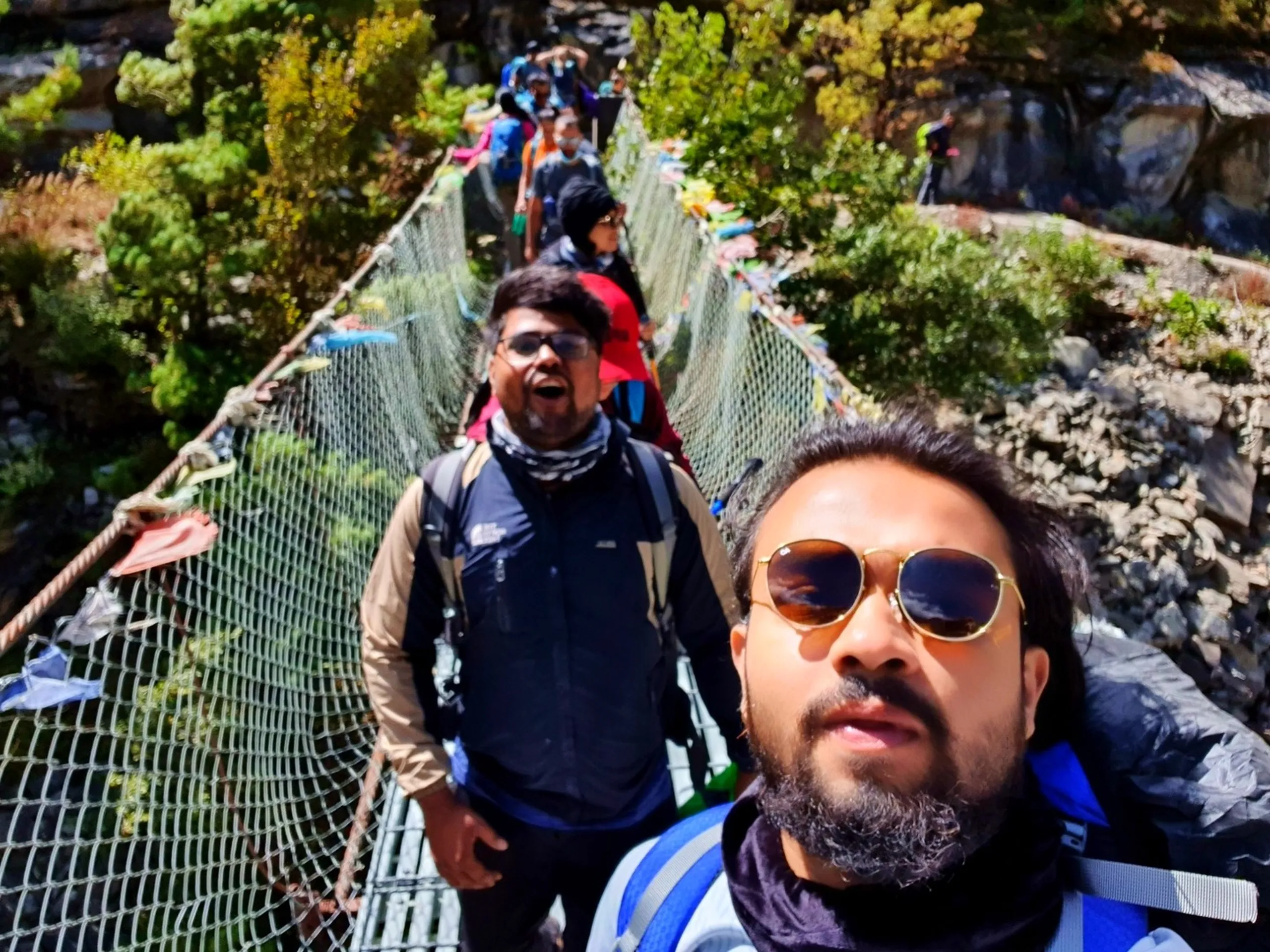 A close-up selfie of two young male Nepali trekkers crossing a prayer flag-adorned suspension bridge on the Annapurna Circuit in Manang, Nepal - the one in the foreground wearing round gold-framed sunglasses, a blue puffer jacket, and a grey neck scarf looking upward - the one behind wearing glasses, laughing openly with mouth agape, dressed in a beige North Face jacket with a checkered scarf - six other trekkers visible crossing the bridge in a line behind them, colorful prayer flags fluttering on both sides - green forest and rocky cliffs visible beyond - a candid and energetic Nepal trekking group bridge selfie, capturing the joy and camaraderie of crossing suspension bridges on the Annapurna Circuit trail to Manang