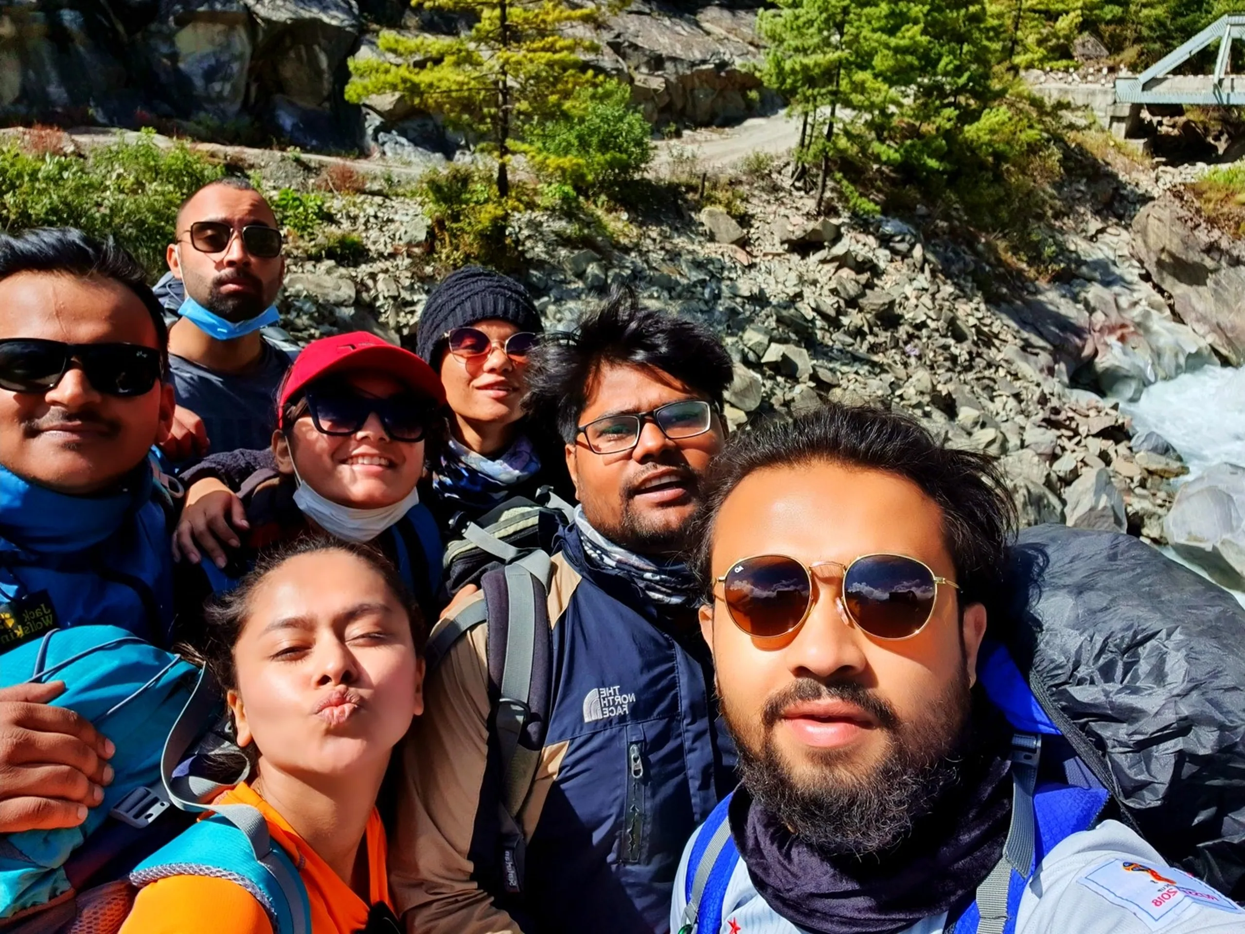 A candid wide group selfie of seven young Nepali trekkers resting beside the turquoise Marsyangdi River near Manang, Nepal - a steel truss bridge and rocky pine-forested cliffside visible behind them - the group in a mix of trekking jackets, caps, sunglasses, and face masks, all crowded close together - one woman in an orange vest making a playful duck-face pout - another woman in a red cap with sunglasses smiling broadly - a man in a North Face jacket in the back - all wearing loaded backpacks - a lively and joyful Nepal trekking group river stop selfie on the Annapurna Circuit, perfectly capturing the family-like bond that forms on a multi-day Himalayan trek in Manang, Nepal