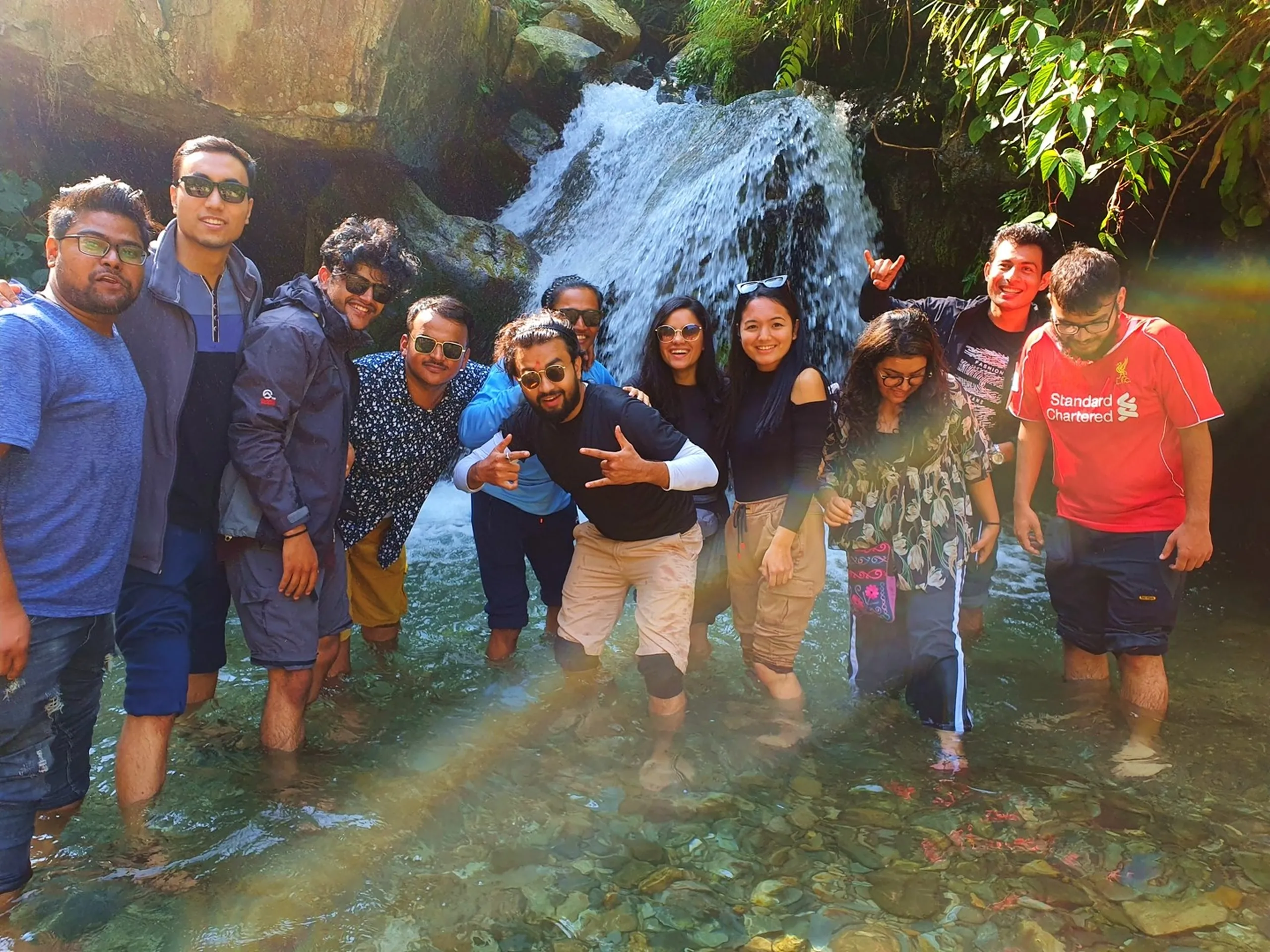 A joyful group of eleven young trekkers standing waist-deep in a clear crystal mountain stream beneath a cascading waterfall surrounded by lush green jungle foliage and mossy boulders in Nepal - laughing, making hand gestures, embracing each other - men and women in casual trekking clothes, some in sunglasses, captured in golden side light - a memorable group waterfall photo on the trail to Lunjung, Lamjung on the Annapurna Circuit, showcasing hidden waterfalls and Nepal trekking adventures with friends