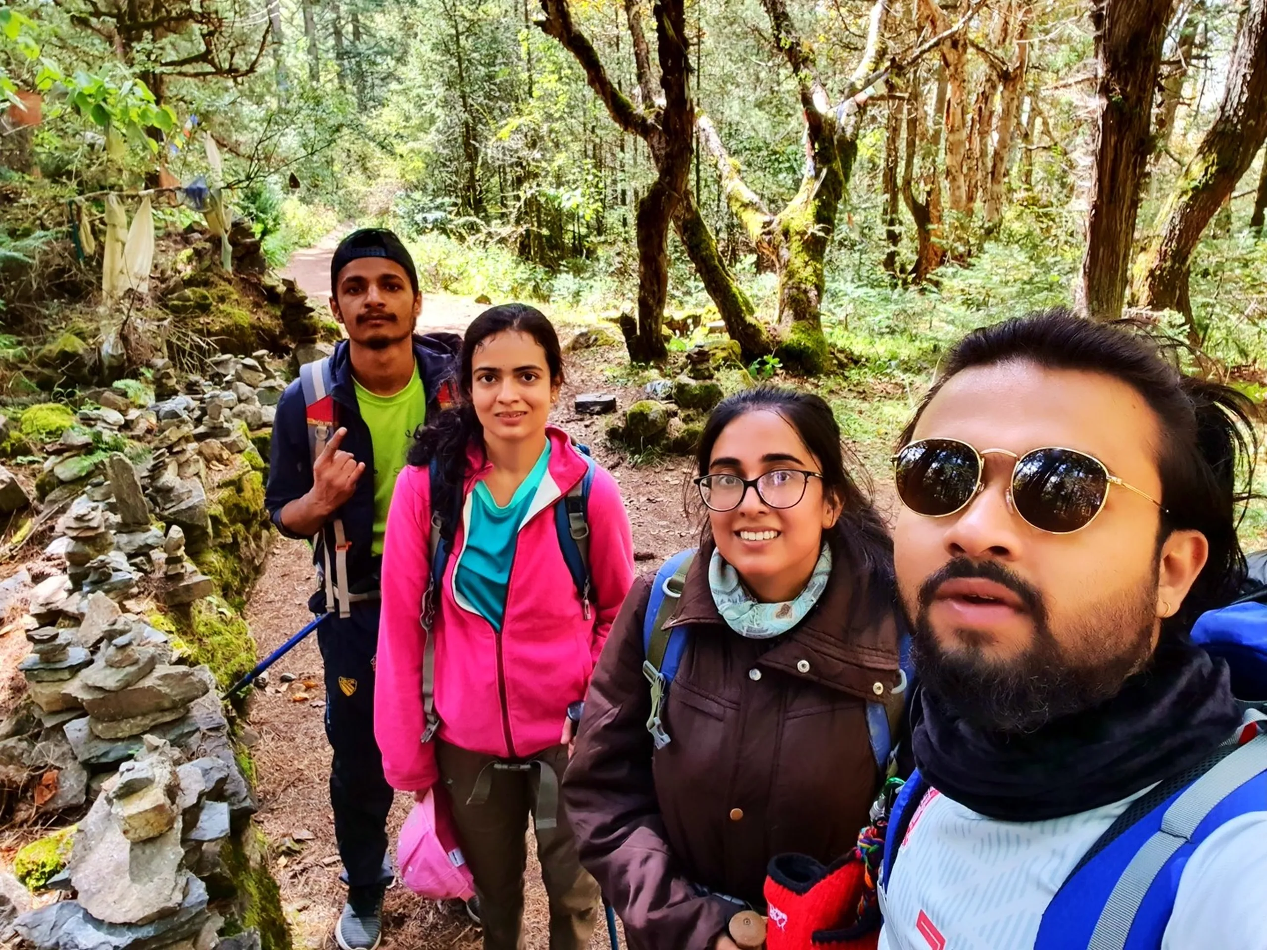 A group selfie of four young Nepali trekkers on a mossy forest trail in Manang, Nepal - the one in the front right wearing round gold-framed sunglasses, a blue jacket, and a colorful scarf - a young woman beside him in a brown jacket and glasses smiling - a young woman in a pink fleece behind them - a young man in a neon green top at the back flashing a rock hand sign - moss-covered stone walls, cairn stacks, and a dappled sunlit Himalayan pine forest with golden and green foliage visible behind - a colorful Tibetan prayer flag barely visible in the tree canopy - a warm and candid Nepal trekking forest trail group selfie on the Annapurna Circuit path to Ice Lake, Manang, capturing the close friendships formed during a Himalayan group trek