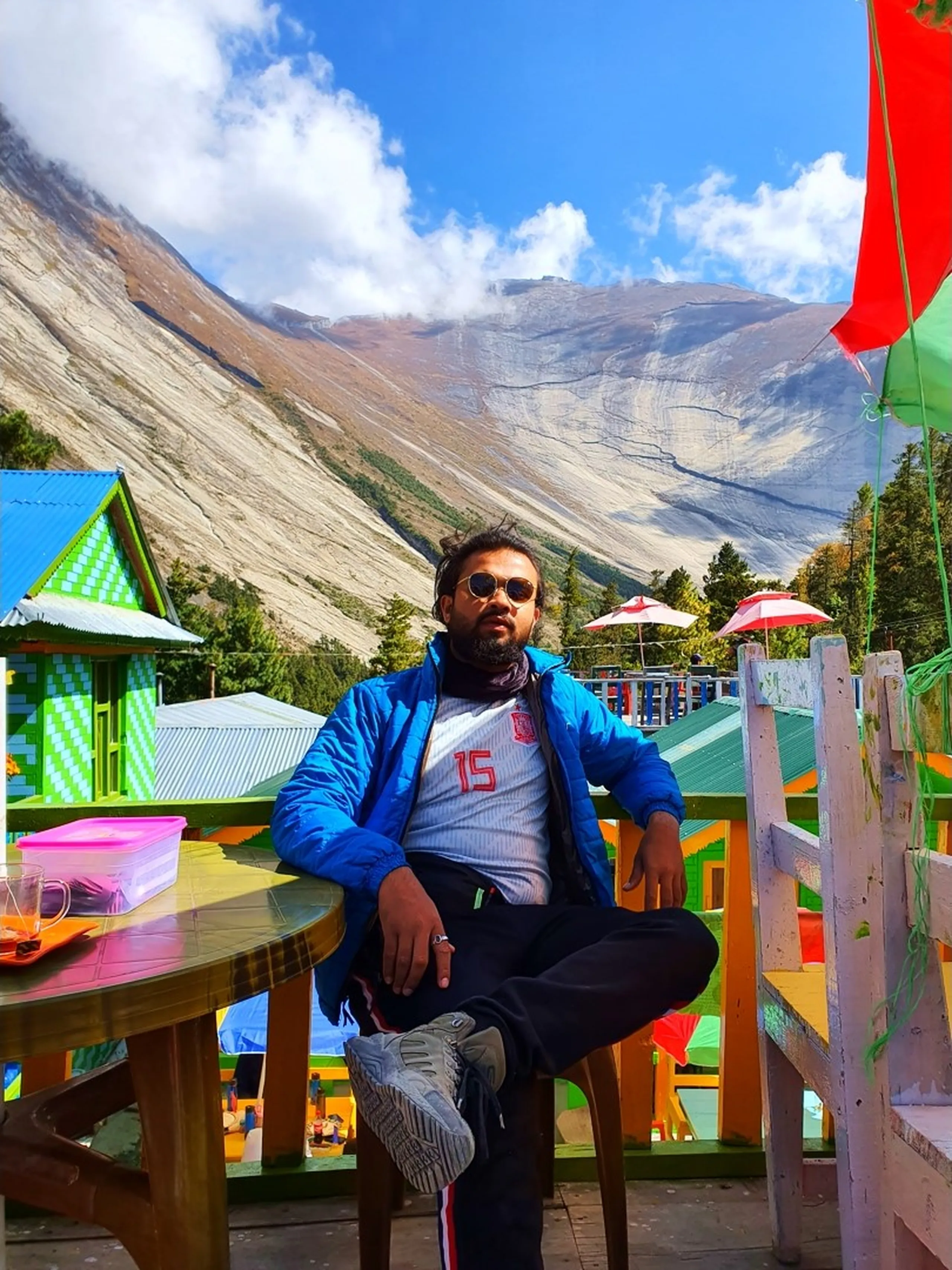 A stylish and relaxed young male Nepali trekker sits in an outdoor chair at a brightly colored teahouse terrace in Manang village, Nepal - wearing a blue puffer jacket open over a white Adidas number 15 football jersey, black track pants with red stripes, and white sneakers - legs casually crossed, one hand resting on the table with a plastic lunch container and a glass nearby - round sunglasses on - a deeply textured, enormous grey and rust-streaked Himalayan mountain face fills the entire background under a blue sky with light cloud - colorful green-and-blue chequerboard and standard painted teahouse buildings visible to the left and behind - red and green umbrellas in the background - a cool and atmospheric Nepal trekking teahouse rest stop portrait in Manang village, Annapurna Circuit