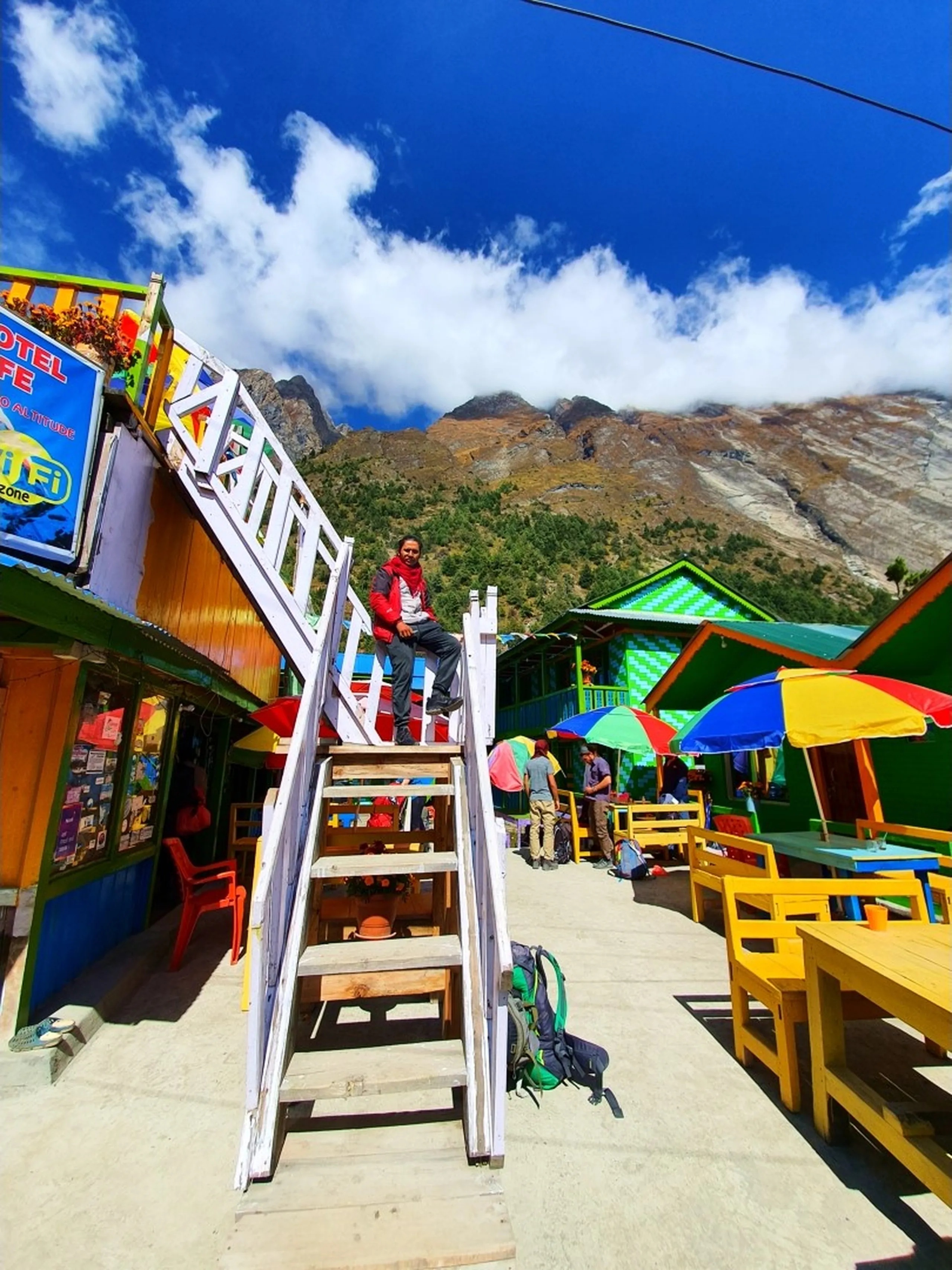 A playful and wide-angle travel photograph of a young male Nepali trekker sitting atop a tall white wooden ladder propped against a teahouse building in vibrant Manang village, Nepal - wearing a red jacket and dark trekking trousers, smiling down at the camera - below him, a bustling and colorful high-altitude teahouse village scene: brightly painted buildings in yellow, green, and blue, outdoor tables with multicolored umbrellas in red, yellow, and blue, a Hotel Cafe WiFi Zone sign, trekkers resting with their gear - dramatic bare grey Himalayan mountain peaks with cloud-wrapped summits rise directly above the village - a fun and vibrant Nepal trekking village photograph in Manang, Annapurna Circuit, capturing the unique energy and color of one of the most iconic teahouse stops on the route to Ice Lake