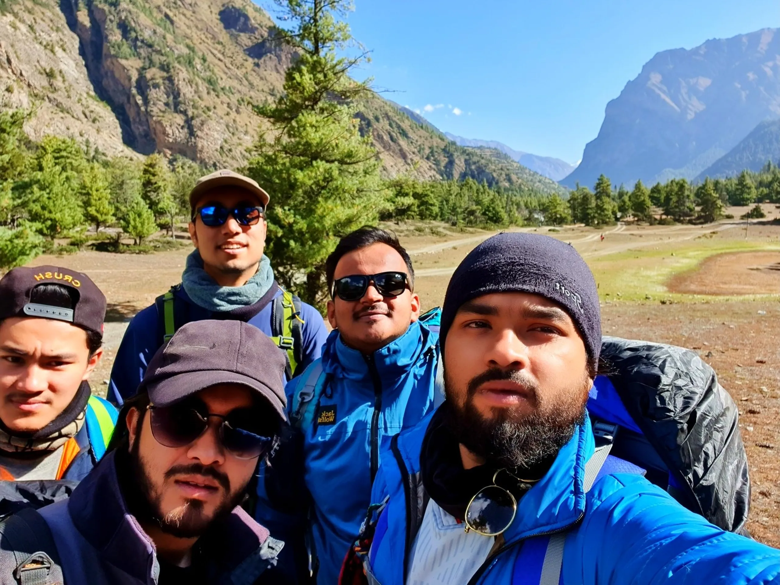 A close-up group selfie of five young male Nepali trekkers in the open Manang valley, Annapurna Circuit, Nepal - the one in the front right wearing a dark blue puffer jacket, wool beanie, and round sunglasses hanging at his collar - another in a purple jacket with sunglasses - one in a brown cap with blue sunglasses - one in a blue Jack Wolfskin jacket - one in a neon green jacket at the far left - behind them, a sweeping Himalayan panorama: open golden alpine meadow, sparse pine trees, layered mountain ridges receding into the blue valley haze - a vibrant and masculine Nepal trekking trail group selfie in the Manang valley, capturing the tight-knit brotherhood formed on the Annapurna Circuit trek to Ice Lake Nepal