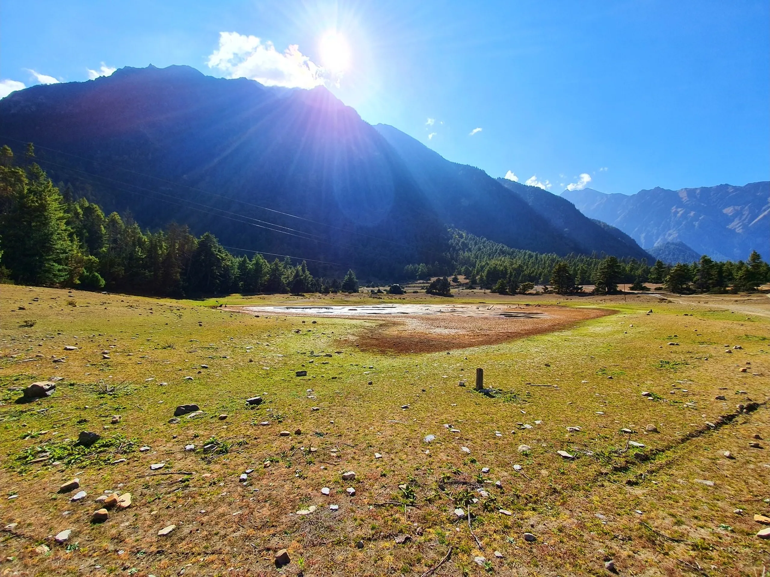 A stunning wide-angle landscape photograph of a high-altitude mountain meadow in the Manang valley on the Annapurna Circuit, Nepal - an open grassy plain with patches of dry golden grass and a shallow seasonal pond or wetland at the center reflecting the sky - scattered pine trees dotting the meadow - the sun blazes dramatically from behind a dark rocky Himalayan peak on the left, sending long golden rays fanning across the deep blue sky - another range of rust and grey mountain ridges visible on the right with patches of pine forest - a lone wooden trail marker post in the foreground - a breathtaking and luminous Nepal high-altitude meadow landscape photograph in Manang, ideal for Annapurna Circuit trekking scenery, Nepal nature photography, and Ice Lake Manang travel blog content