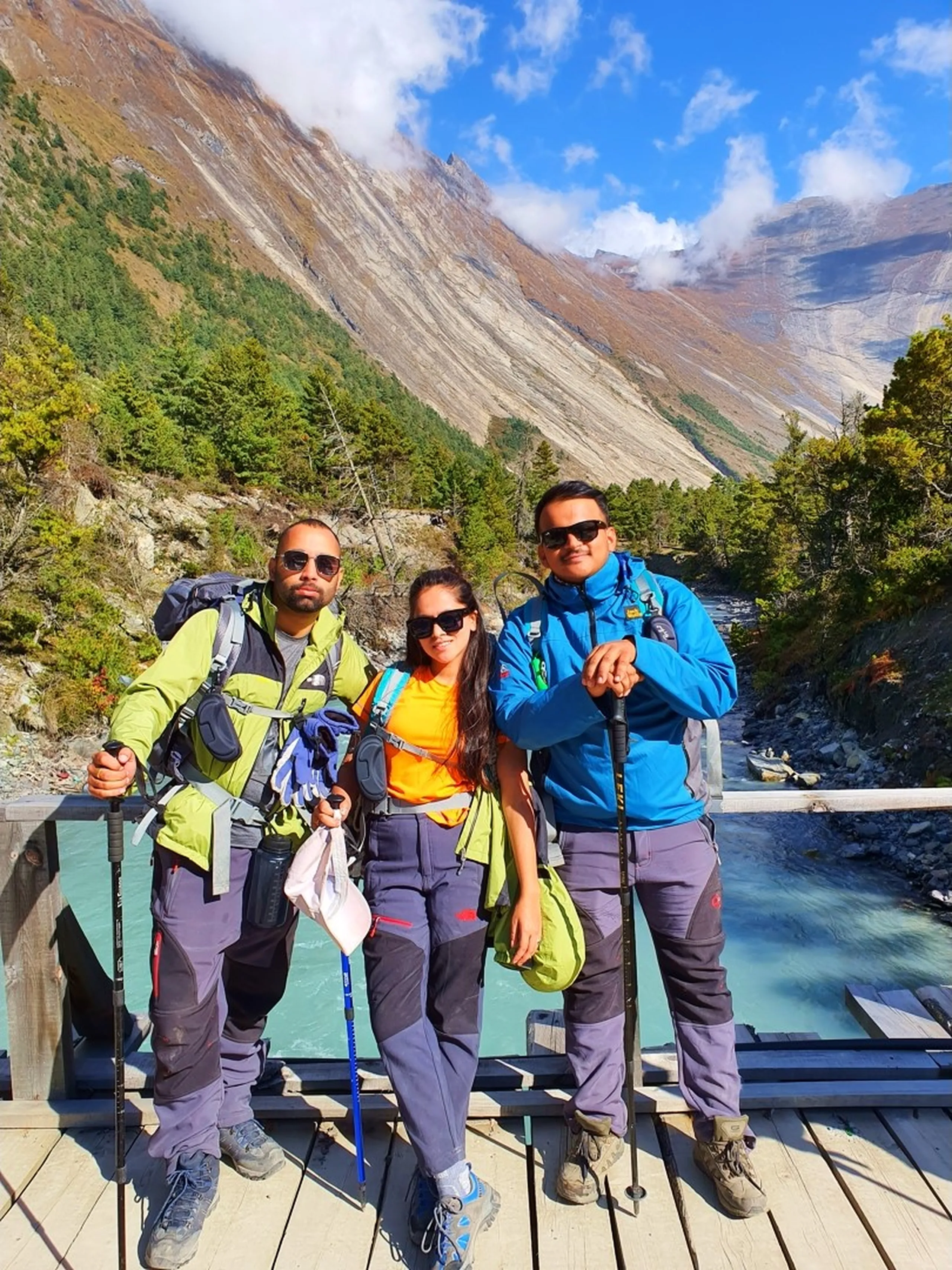 Three young Nepali trekkers pose together on a wide wooden footbridge over the brilliantly turquoise Marsyangdi River in Manang, Nepal - a young man on the left in a neon green-yellow jacket with dark sunglasses and trekking pole - a young woman in the center wearing an orange tee, blue trekking trousers, and dark sunglasses - a young man on the right in a bright blue Jack Wolfskin jacket, sunglasses, and trekking pole - all three leaning against the wooden railing with the stunning glacial river flowing brilliantly below - a dramatic grey and rust Himalayan valley wall with pine forest and low cloud visible behind - a vibrant and colorful Nepal trekking bridge portrait near Manang on the Annapurna Circuit, showcasing friendship, adventure, and the iconic turquoise rivers of the Himalayan trekking trail