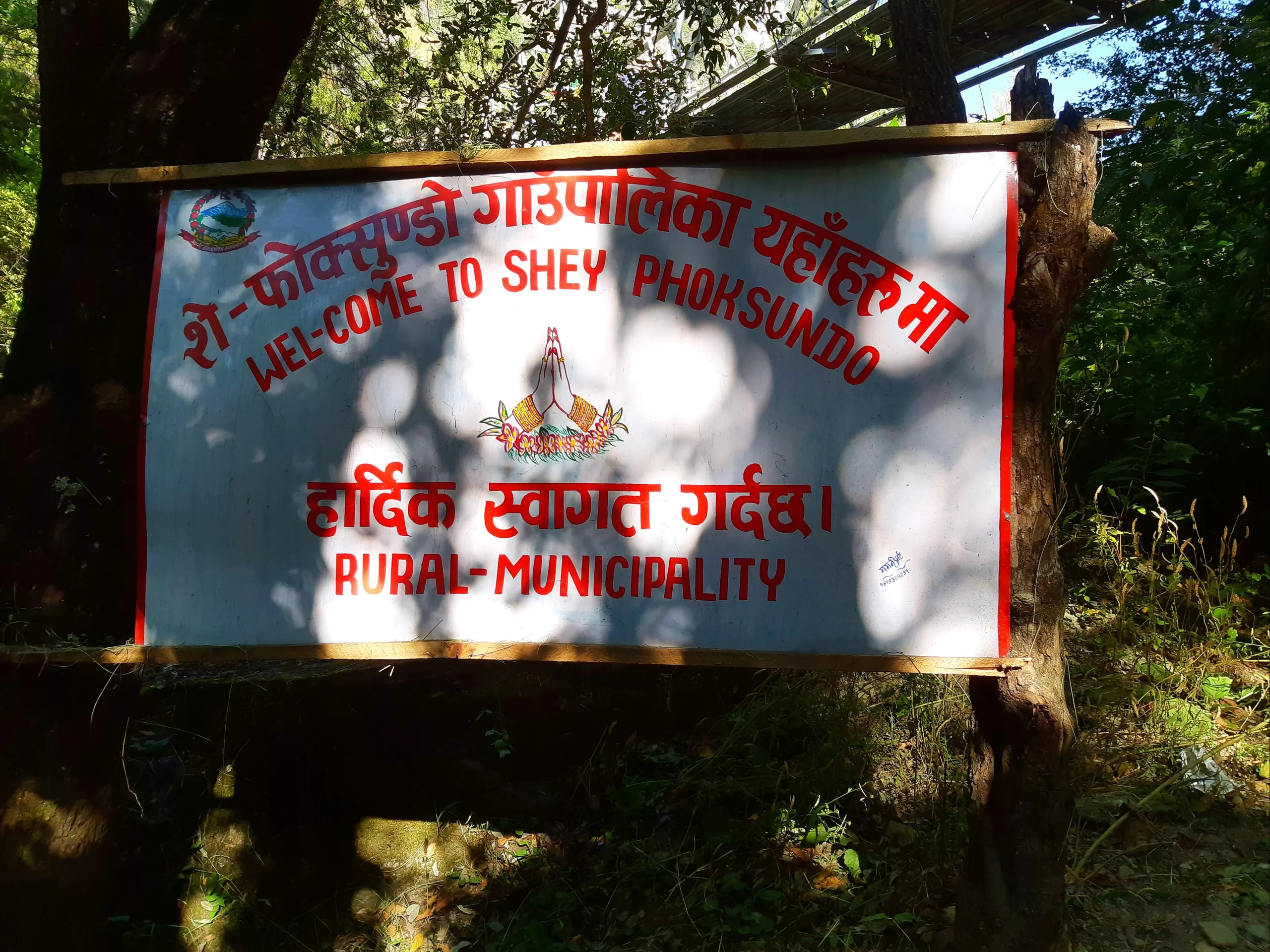 A rectangular welcome signboard mounted on two wooden posts at the edge of a forest path. The board has a white background with a bold red rectangular border. The text reads in large letters: "Welcome to SHEY PHOKSUNDO" in English, with Nepali text above 