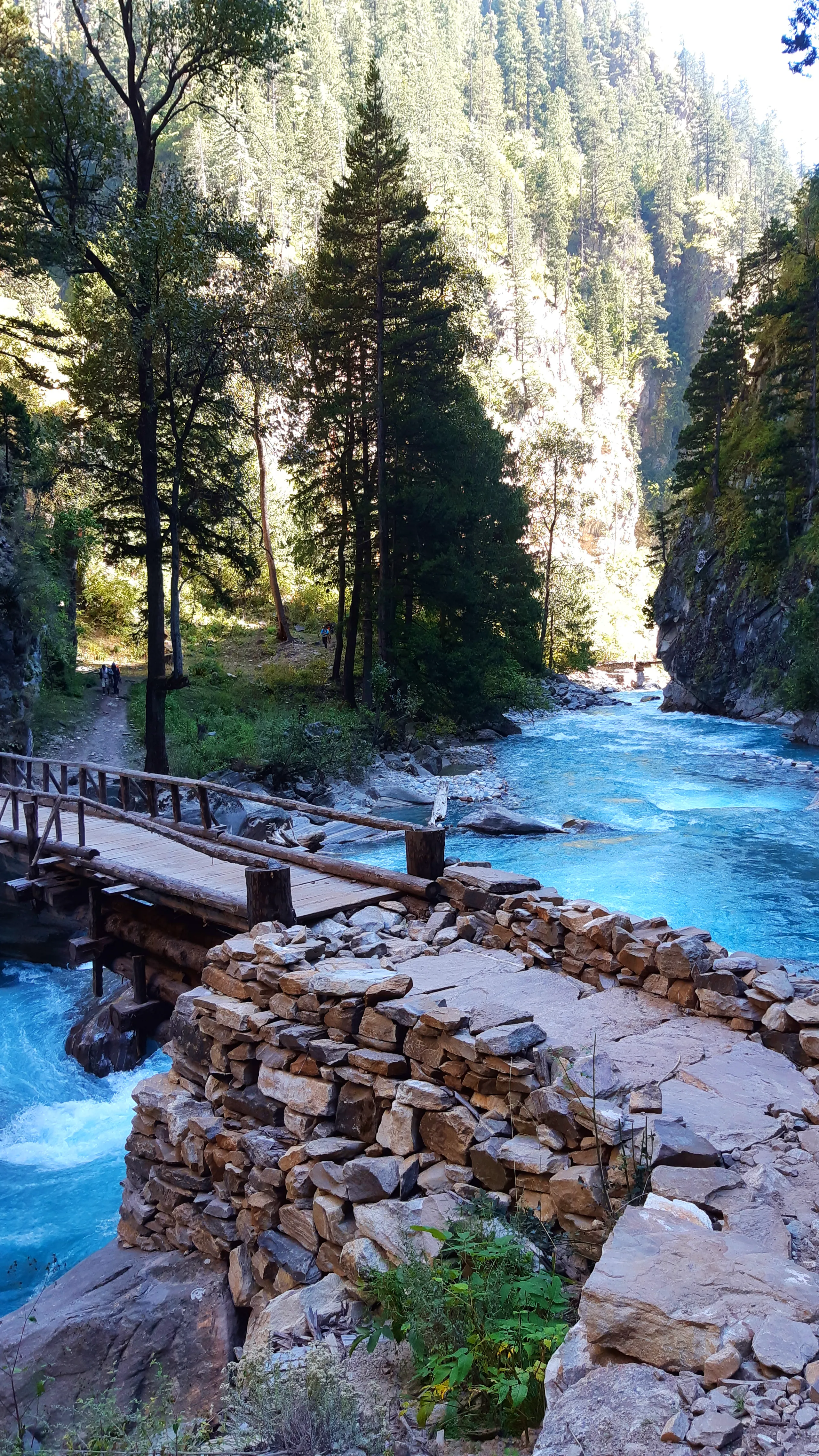 A serene yet dramatic mountain gorge scene. In the foreground, a carefully constructed dry-stone wall made of flat, tightly-fitted grey stones rises from the rocky ground. Beyond it, a wooden plank-and-railing bridge crosses a gorge over a vividly turquoi