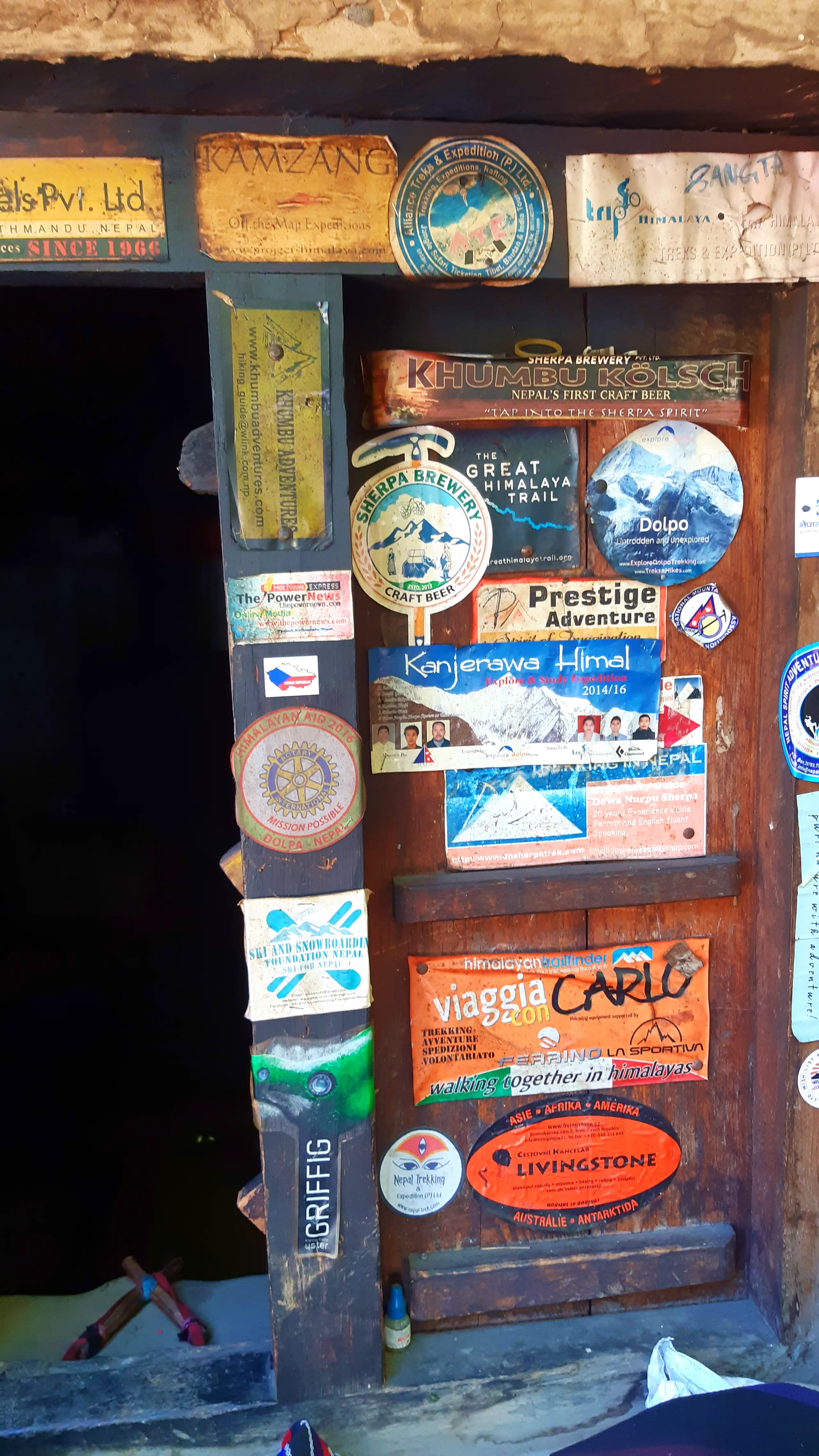 A rustic wooden door of a remote teahouse or lodge, completely covered in an eclectic collage of stickers, badges, and patches left by trekkers and adventurers over many years. The door is dark-stained wood set within a weathered wooden frame. Among the m