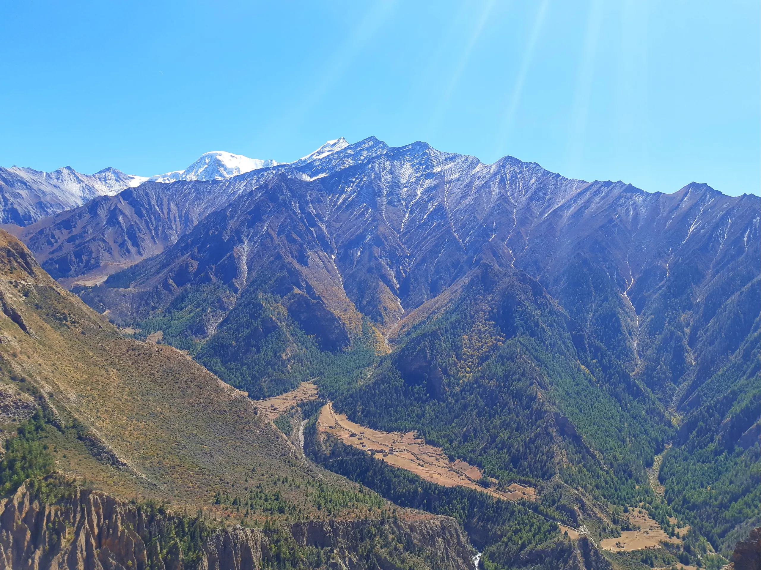 A sweeping panoramic view of a high Himalayan mountain range. In the far background, snow-capped peaks gleam brilliant white against a deep cobalt-blue sky, their summits sharp and jagged. In the middle distance, steep ridgelines are blanketed in dark gre