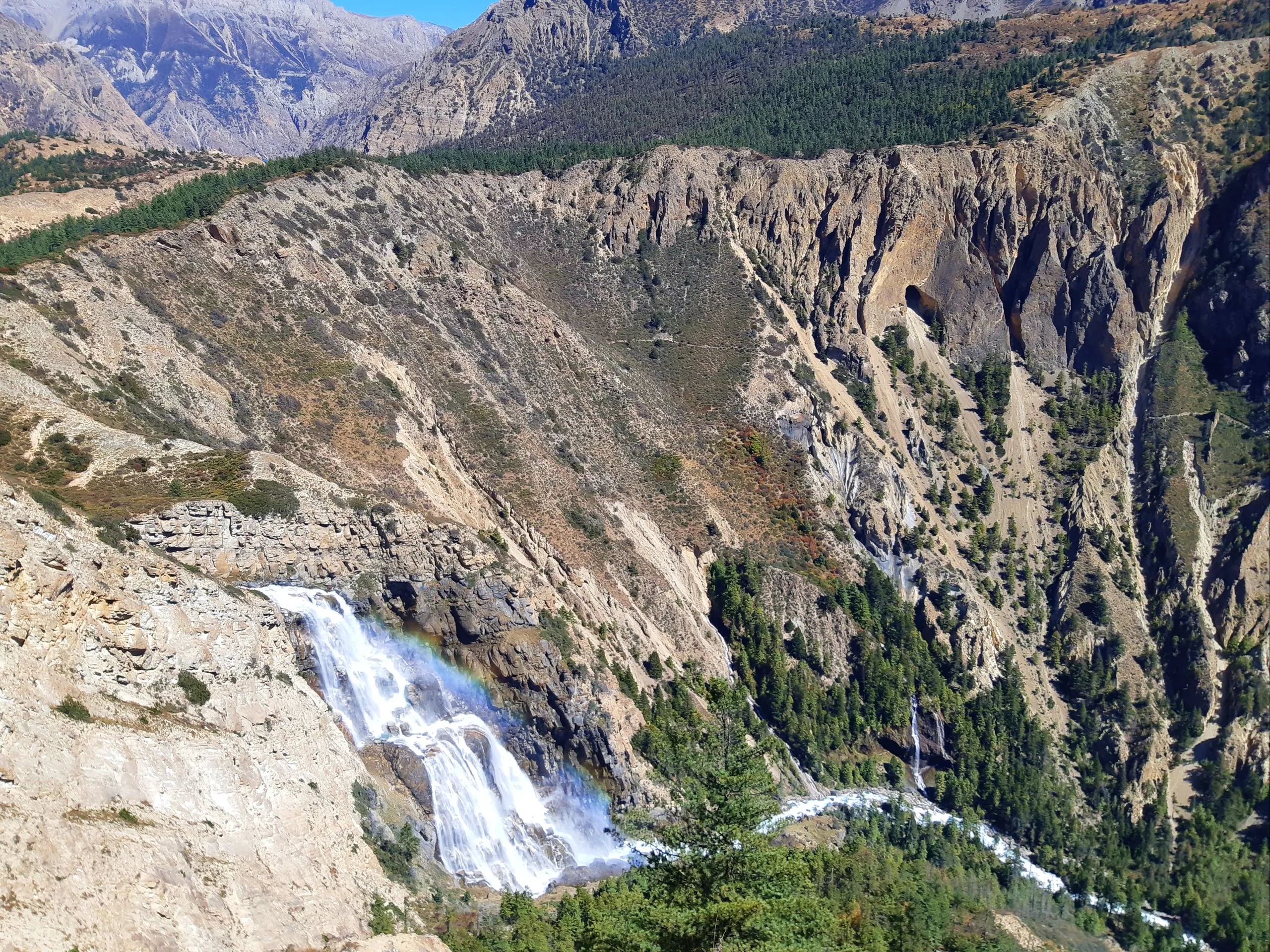 A powerful waterfall plunges dramatically down a sheer, barren rocky cliff face in a remote mountain canyon. The white water falls in wide, fan-like streams across the broad rock face before crashing and dispersing at the base. The surrounding terrain is 