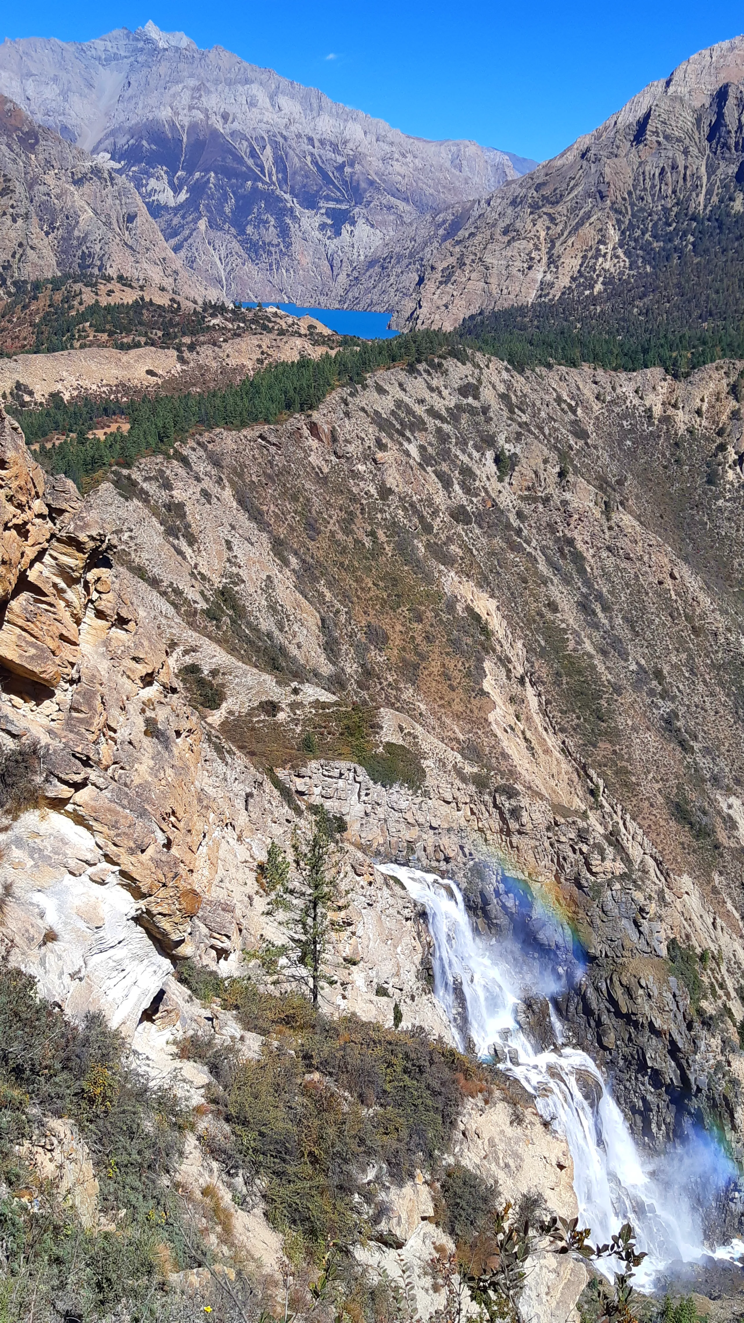 A dramatic, vertically-composed mountain landscape showing a large waterfall tumbling down rugged rocky terrain in the foreground, with a breathtaking surprise in the background: far above and behind, a strikingly beautiful deep cobalt-blue alpine lake - 
