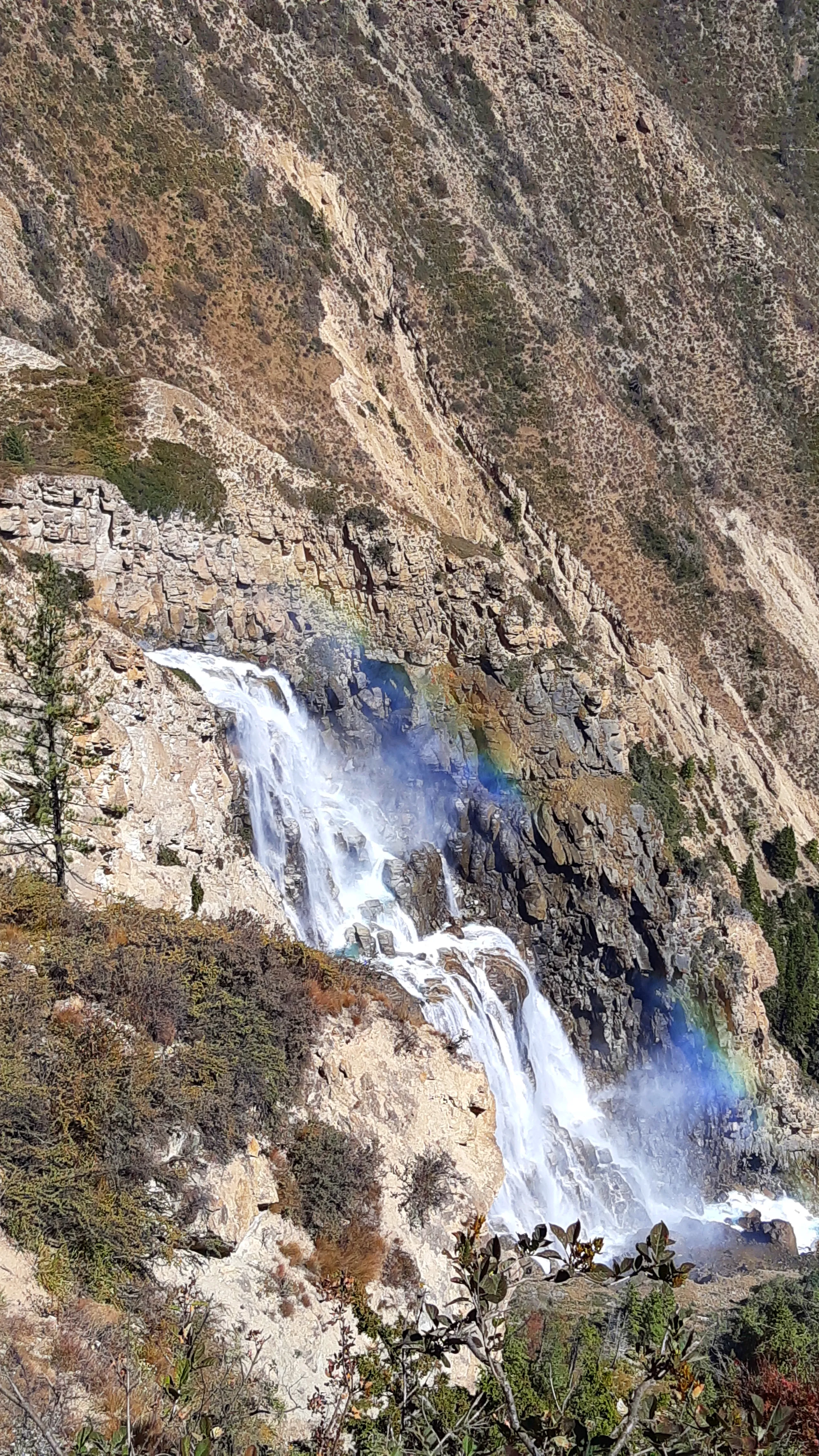 A close-up view of a powerful multi-tiered waterfall cascading down a steep, dry, rocky mountainside. The water crashes and sprays in thick white torrents over the rugged rock face. At the base of the main falls, a vivid rainbow arcs through the mist - it