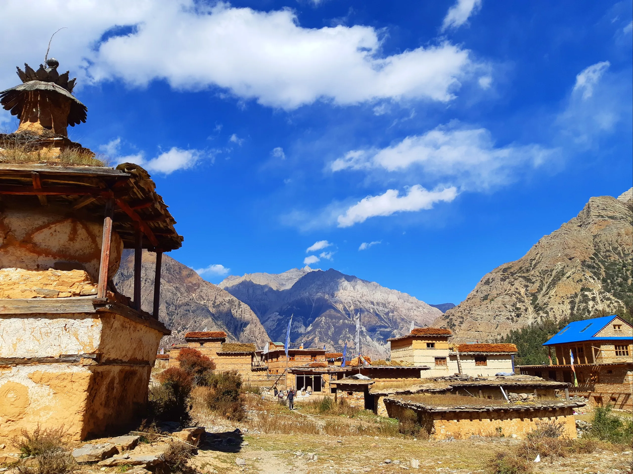 A traditional high-altitude Himalayan village bathed in bright midday light. In the foreground to the left stands a well-worn stone chorten (Buddhist stupa) - a tiered, ornamental religious monument with a square base, tapering body, and decorative top pi
