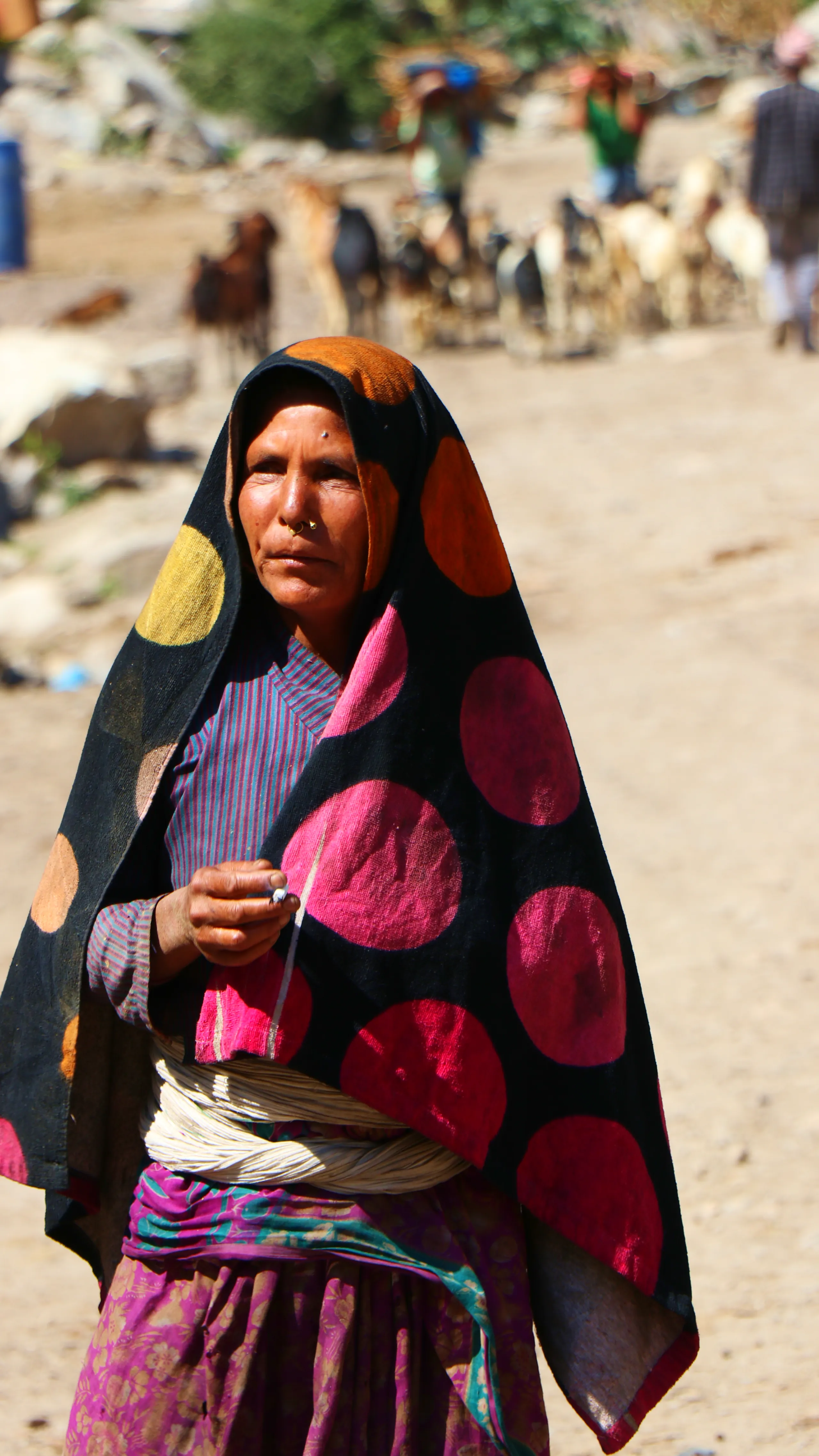 A striking close-up portrait of an older woman standing outdoors in a dusty, open setting. She is draped in a large, bold black shawl or wrap that covers her head and shoulders, decorated with oversized polka dots in vivid pink, magenta, orange-yellow, an