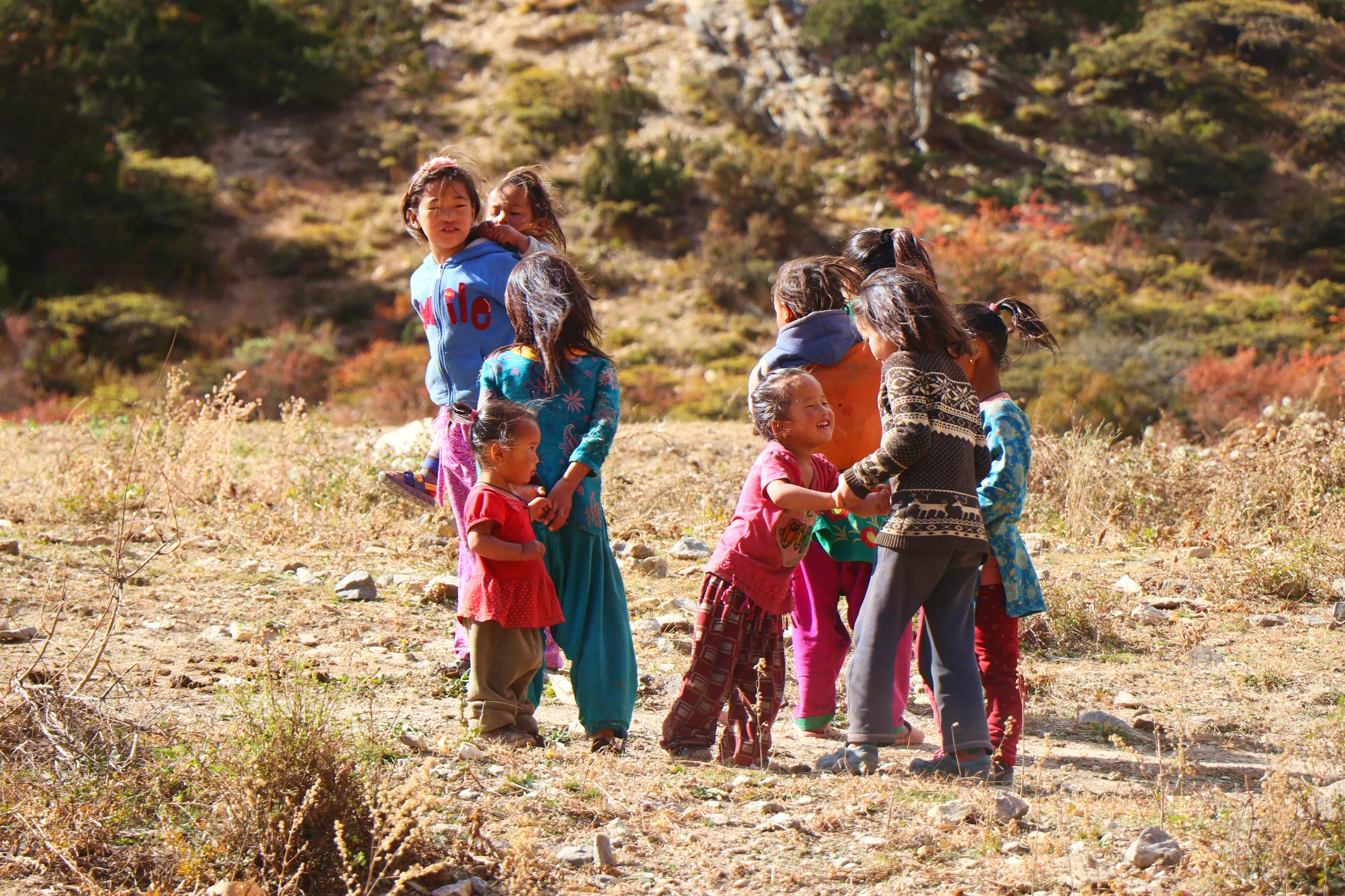 A candid group shot of six or seven children of varying ages gathered on a dry, open hillside. The landscape around them is arid - parched golden-brown earth and dry grass, with sparse red and orange autumn shrubs on the slope behind. The children are dre