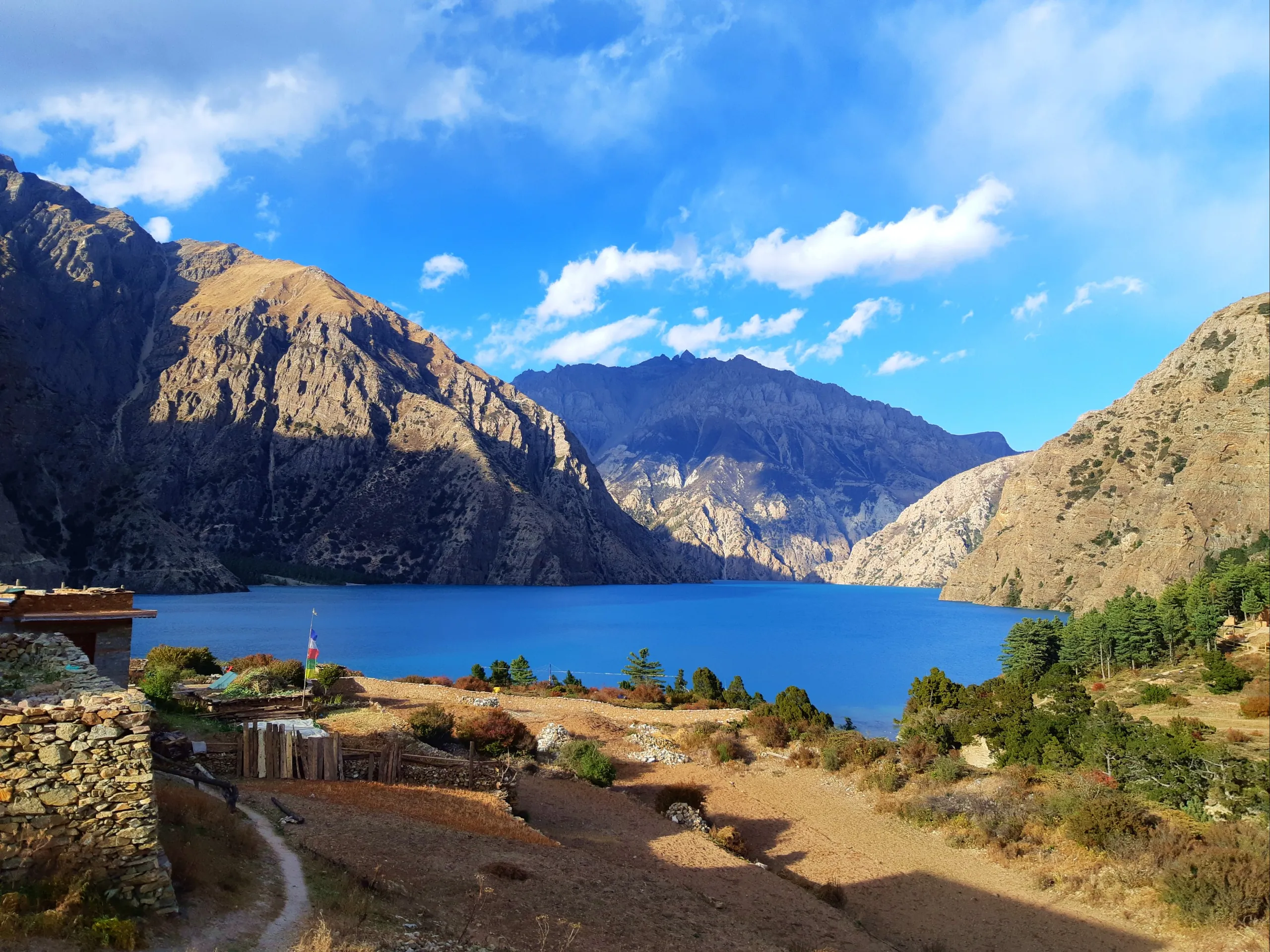 A wide-angle view of a stunning alpine lake - almost certainly Phoksundo Lake in Nepal's Dolpa district - set within a bowl of massive, dramatically steep rocky mountains. The water is an intense, vivid turquoise-blue, completely calm, reflecting the moun