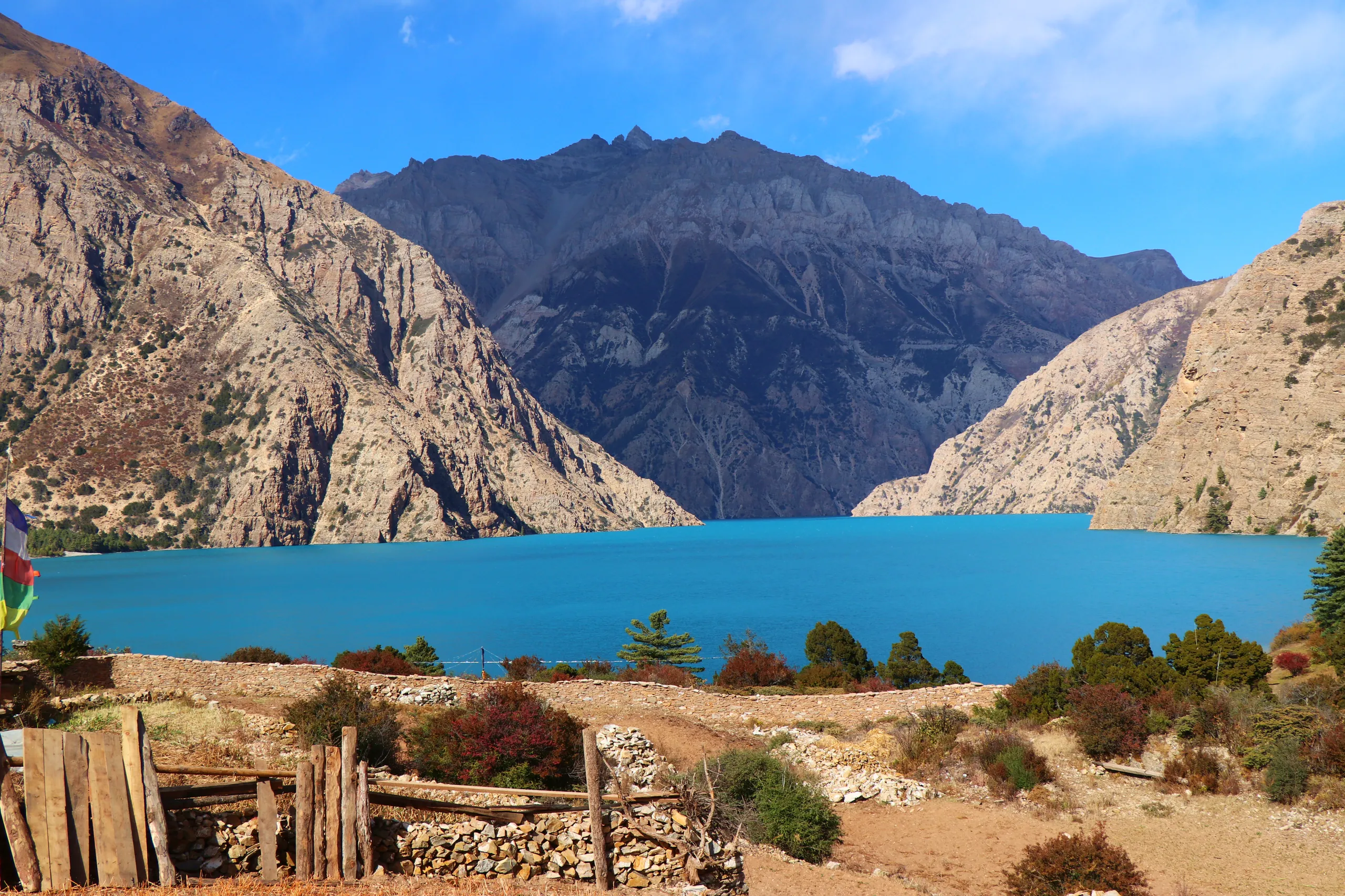Another breathtaking view of Phoksundo Lake, its water an extraordinarily vivid teal-turquoise that seems almost artificially saturated. The lake stretches wide across the center of the frame, enclosed on three sides by massive, sheer rocky mountain walls