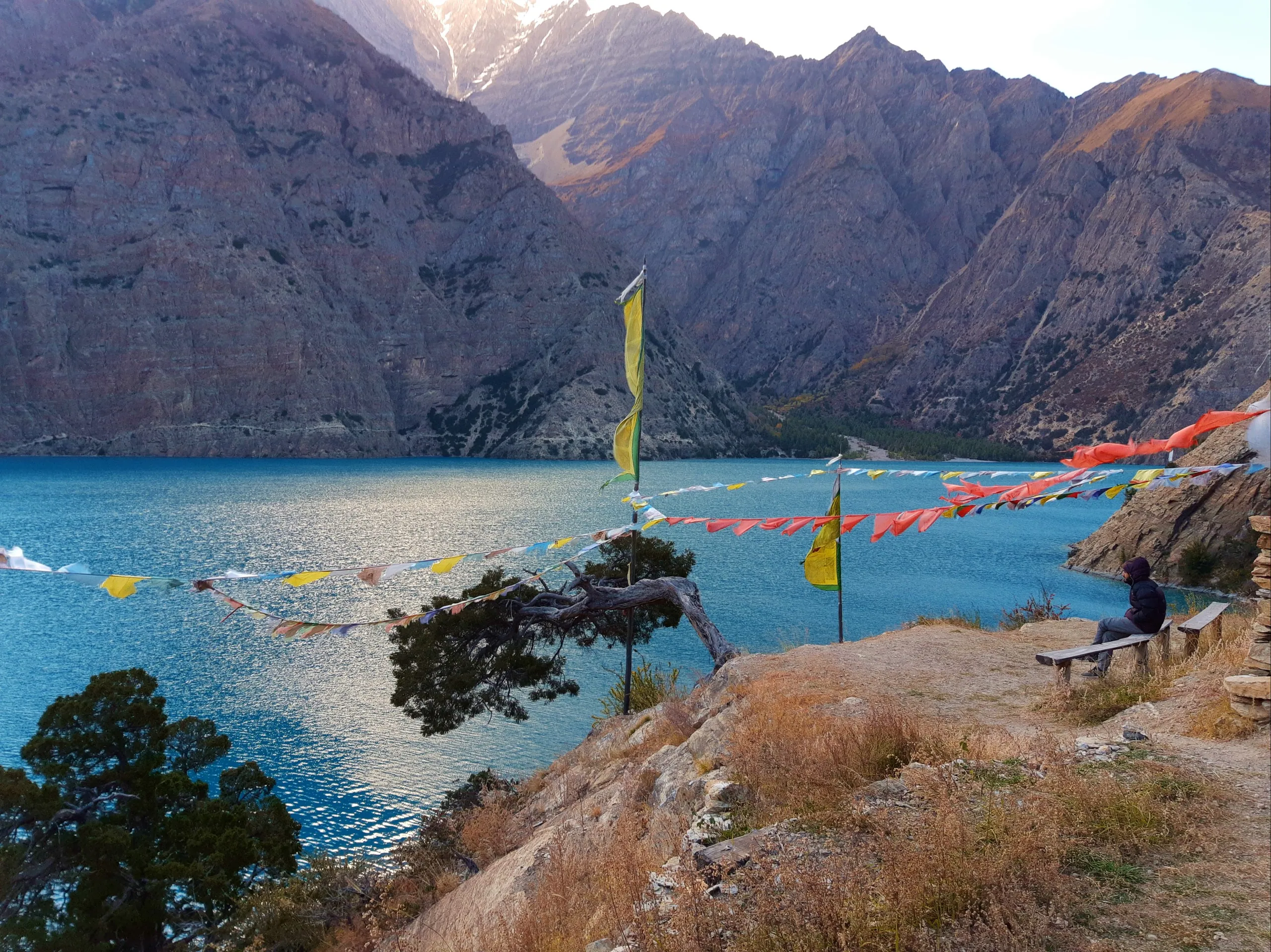 A serene lakeside scene at Phoksundo Lake, captured in the warm light of late afternoon. In the foreground, colorful Buddhist prayer flags - in the traditional five colors of yellow, red, green, white, and blue, along with orange - are strung on lines bet