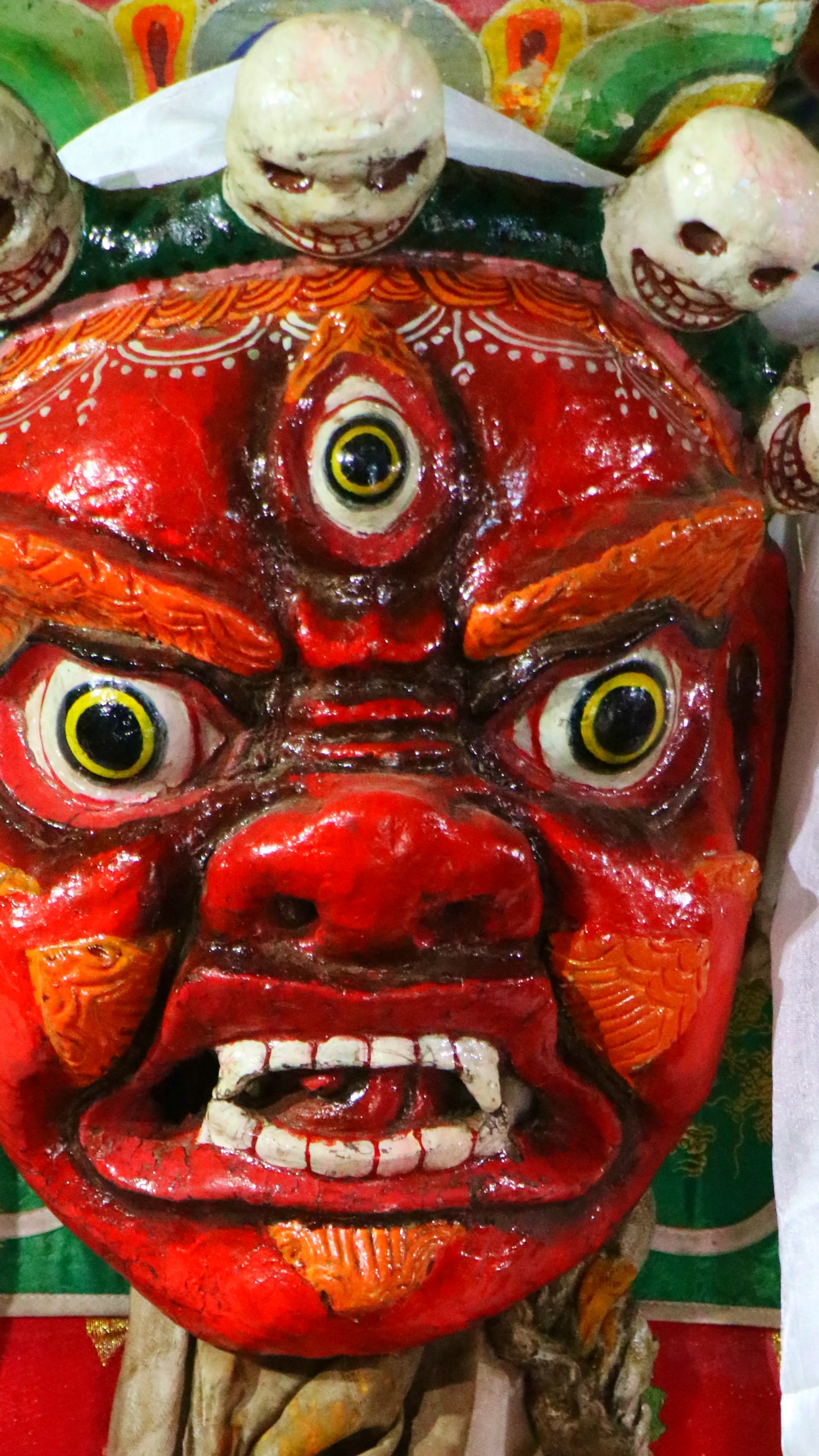 An extreme close-up of a large, fearsome Tibetan Buddhist wrathful deity mask or statue - almost certainly a Mahakala or similar protective dharmapala (dharma guardian). The face is entirely painted in glossy, vivid red. It bears three eyes - two large, b