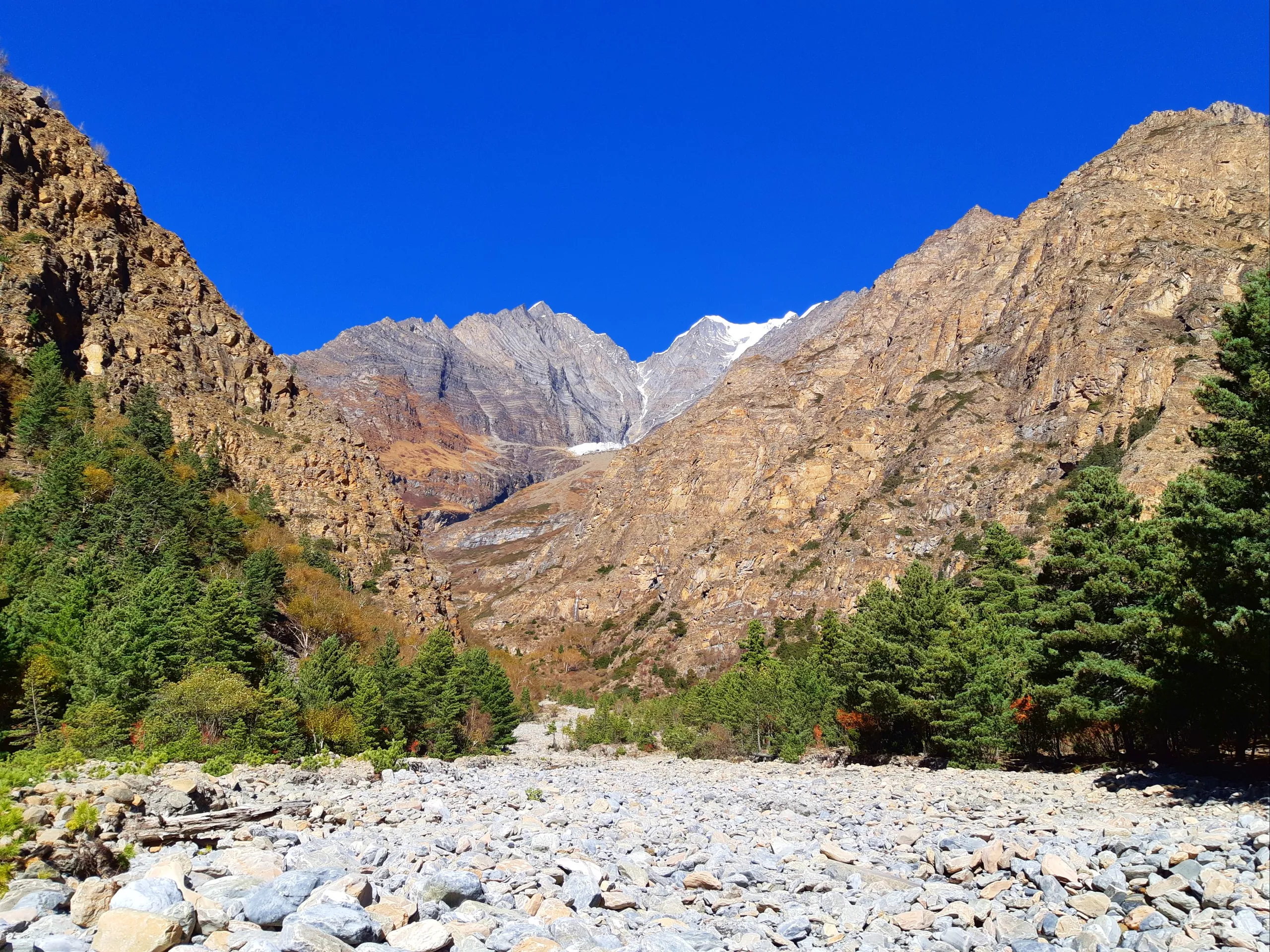 A wide glacial river valley with a completely dry, broad, flat riverbed carpeted entirely in smooth, rounded grey and white boulders and river stones - the classic look of a glacial moraine or seasonal riverbed. Not a drop of water is visible; the river h