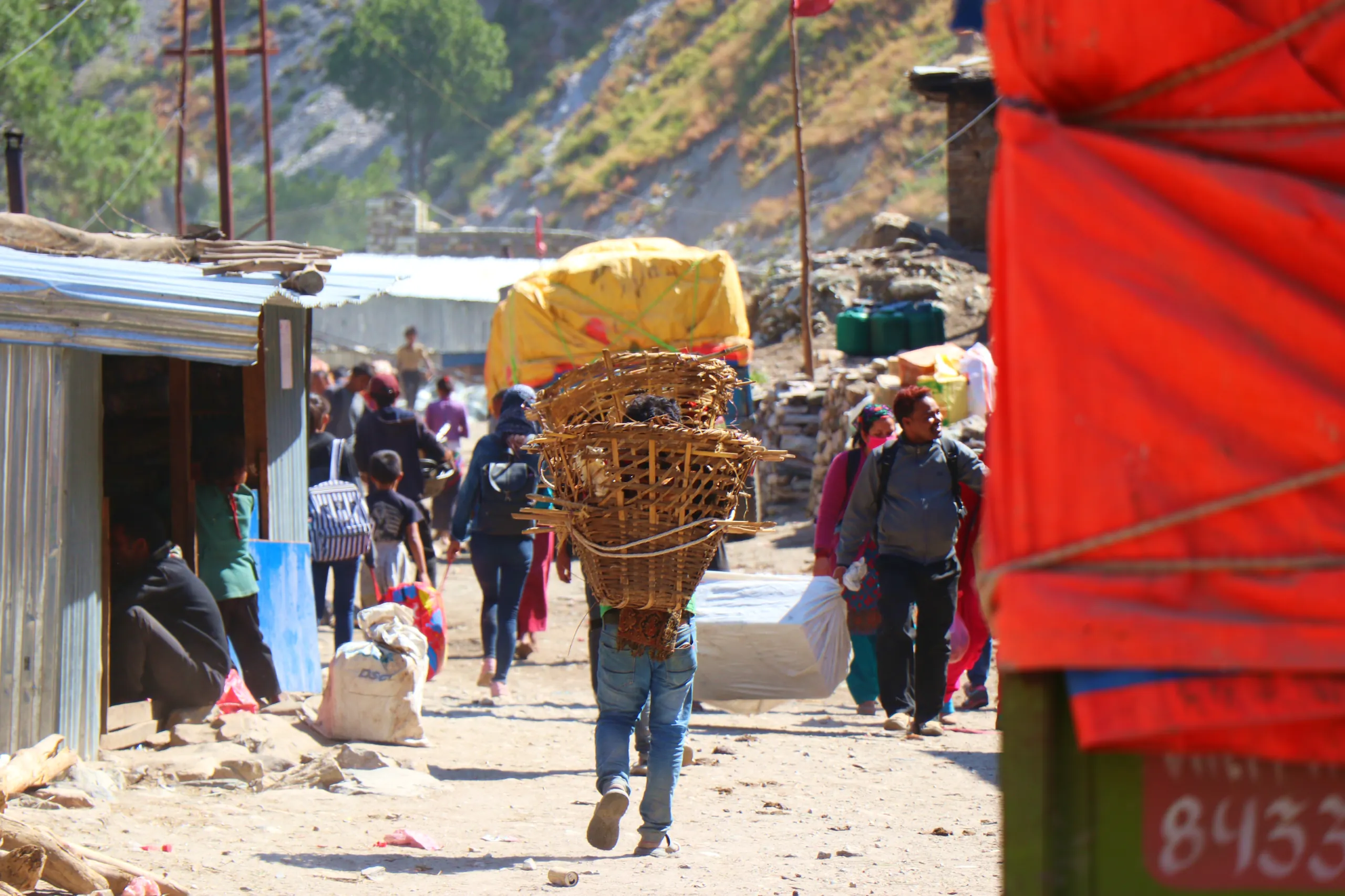 A busy, dusty village market path through what appears to be a remote Himalayan bazaar. In the center of the frame, a solitary porter - a small figure against their enormous load - walks forward carrying an impossibly tall stack of goods: a large bright y