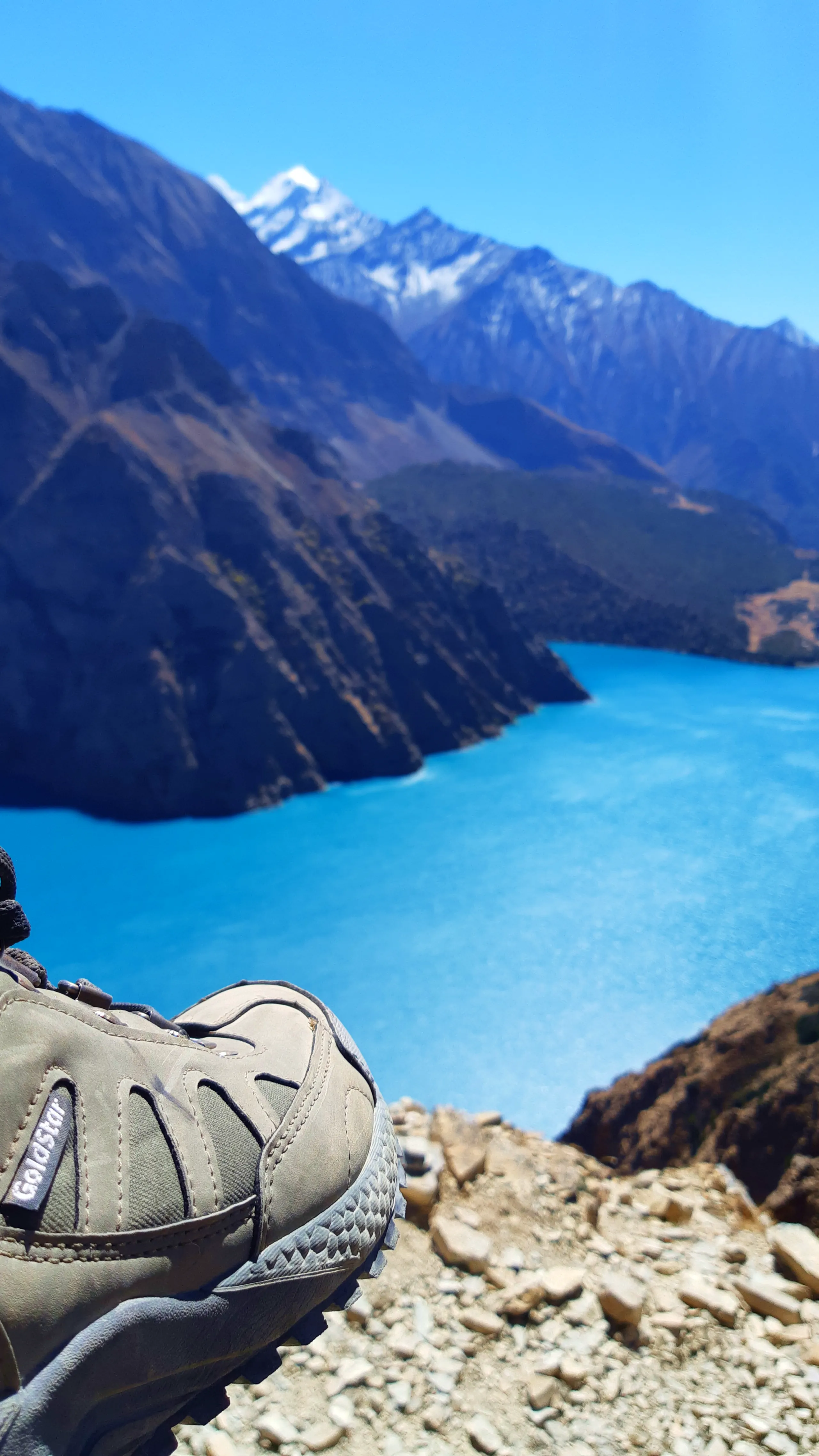 A personal, playful trekker's photograph. In the sharp foreground, a well-worn hiking shoe or trail sandal - grey-olive green, bearing the Merrell brand logo on the side, its sole dusty and roughened from hard use - rests propped on a rocky ledge, as thou