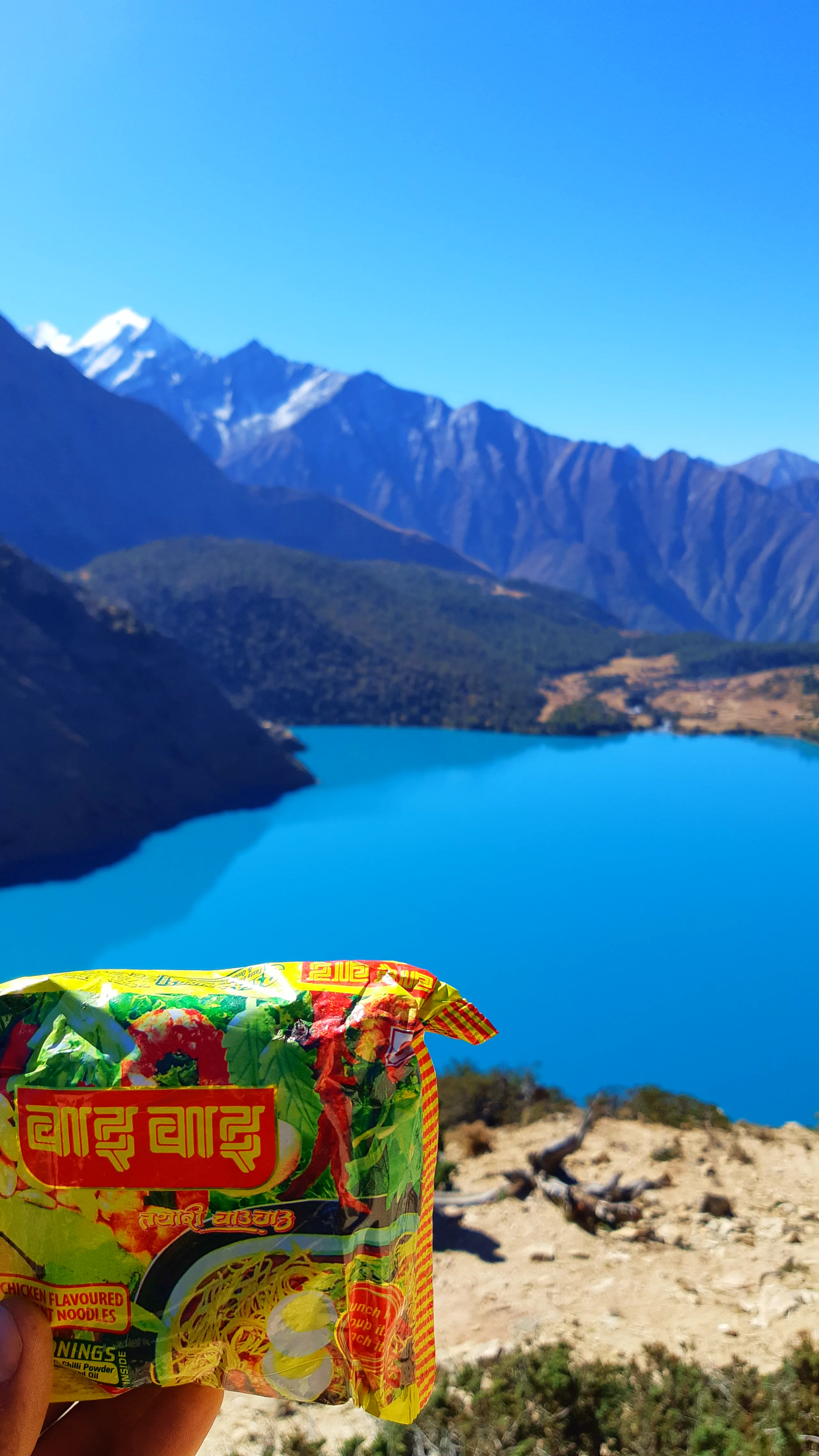 A deeply human and humorous travel photograph. A hand extends into the bottom of the frame, holding up a brightly colored packet of Wai Wai instant noodles - Nepal's beloved, ubiquitous snack food - directly in front of the camera lens. The packaging is v