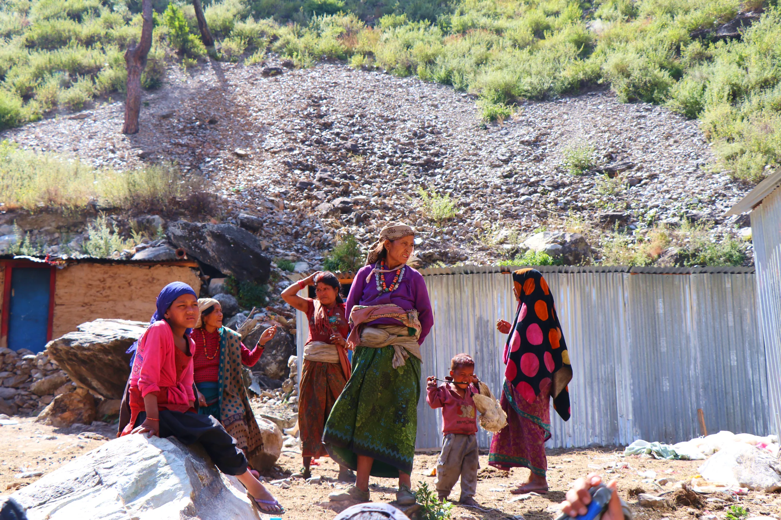 A candid, unposed scene at what appears to be a temporary or semi-permanent settlement on rocky, bare mountain terrain. Three or four women of varying ages stand or move in the mid-ground. One wears a bright pink top with a green patterned skirt; another 