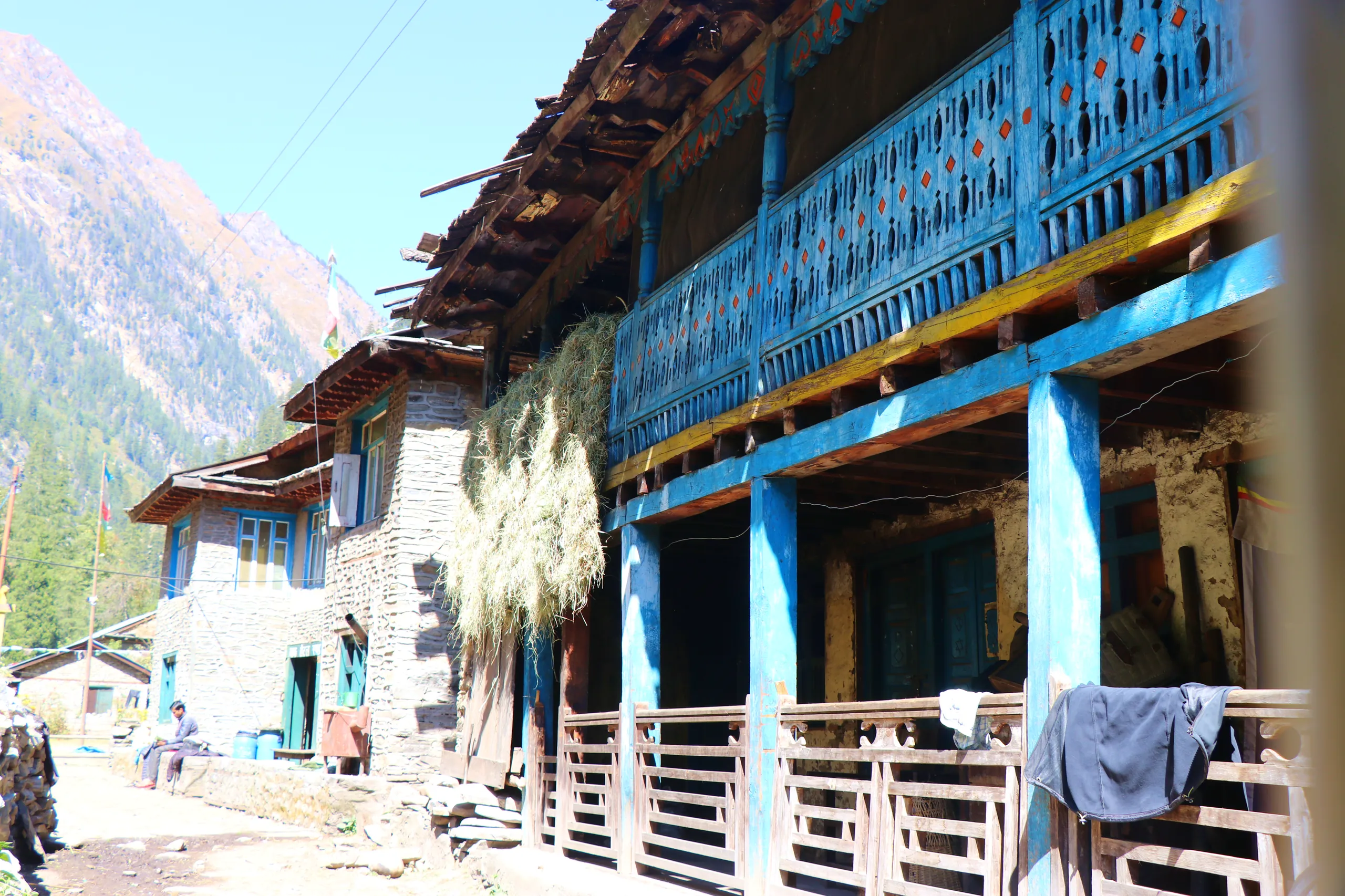 A mountain village street scene centered on a striking, multi-story building whose entire upper veranda is painted in bold turquoise-blue - the color vivid and eye-catching against the otherwise earthy tones of the surroundings. The upper floor has a wide