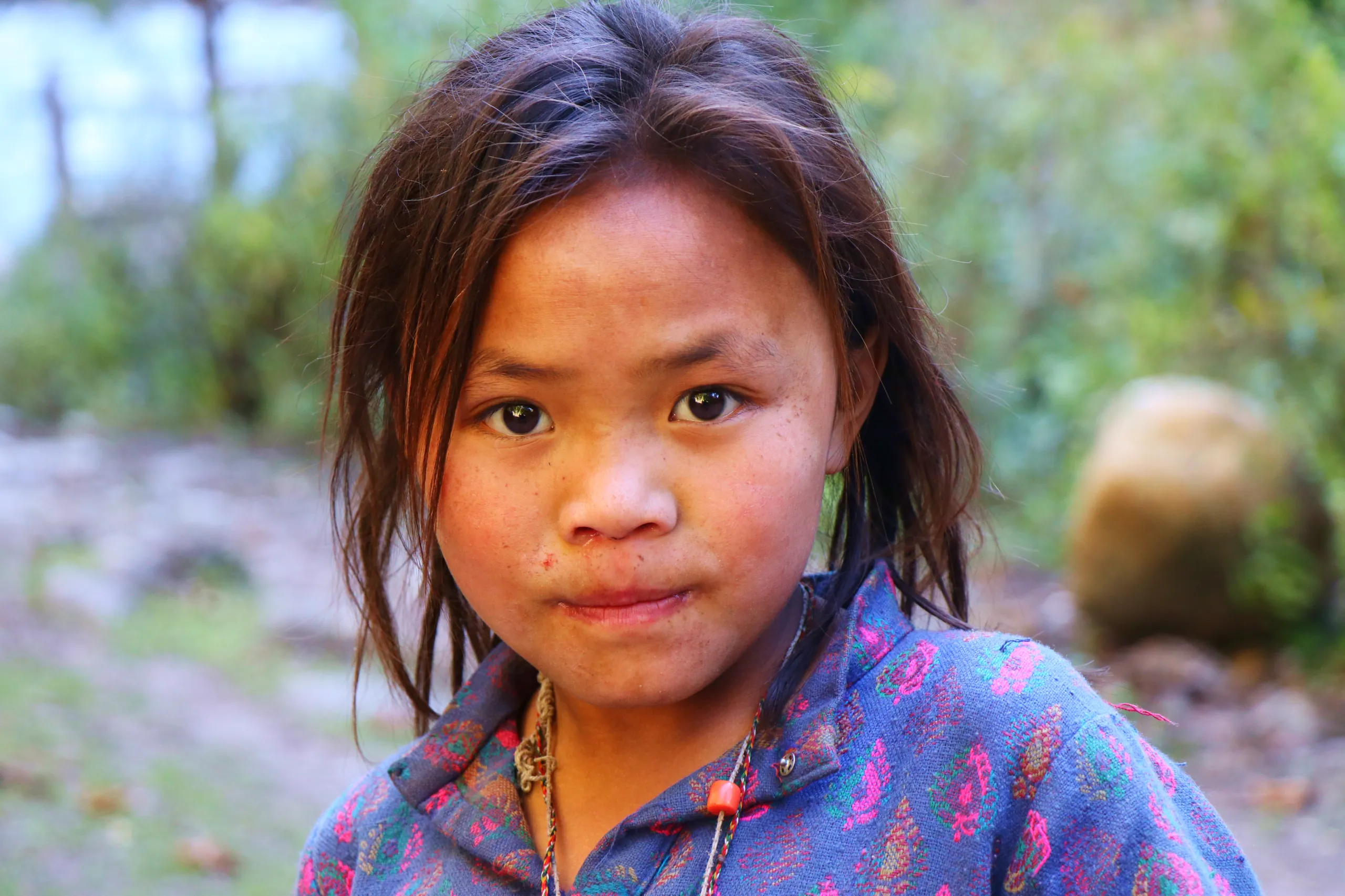 A quiet, close-up portrait of a young girl, approximately 8-10 years old, with South Asian or Himalayan features. Her dark hair falls loosely around her face in slight disarray. She looks directly and steadily into the camera with large, dark, calm eyes -