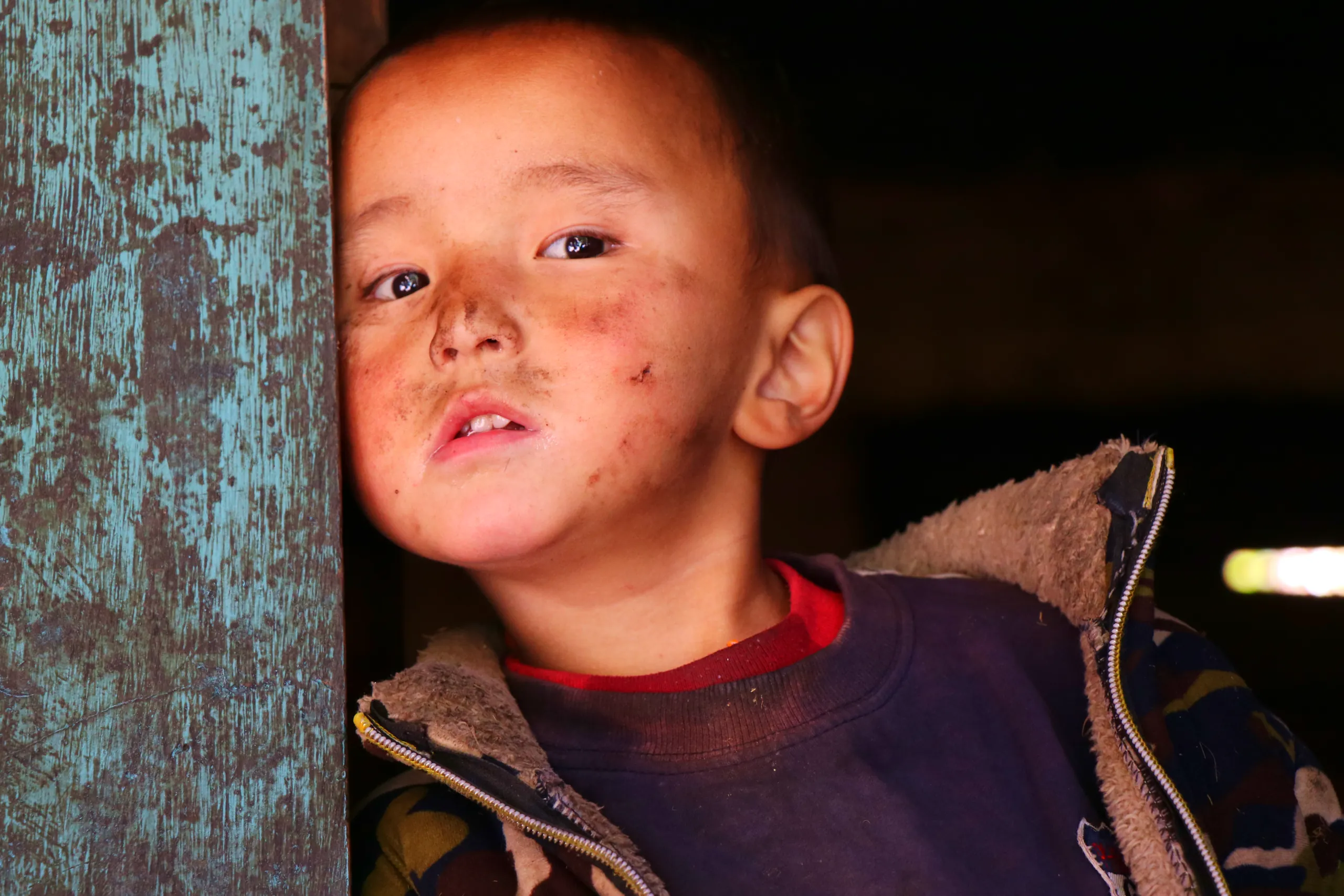 A close, intimate portrait of a young boy, approximately 6-8 years old, leaning gently against a door frame or post covered in peeling, weathered turquoise-green paint - the flaking texture adding richness to the image's edges. The boy has short, dark hai