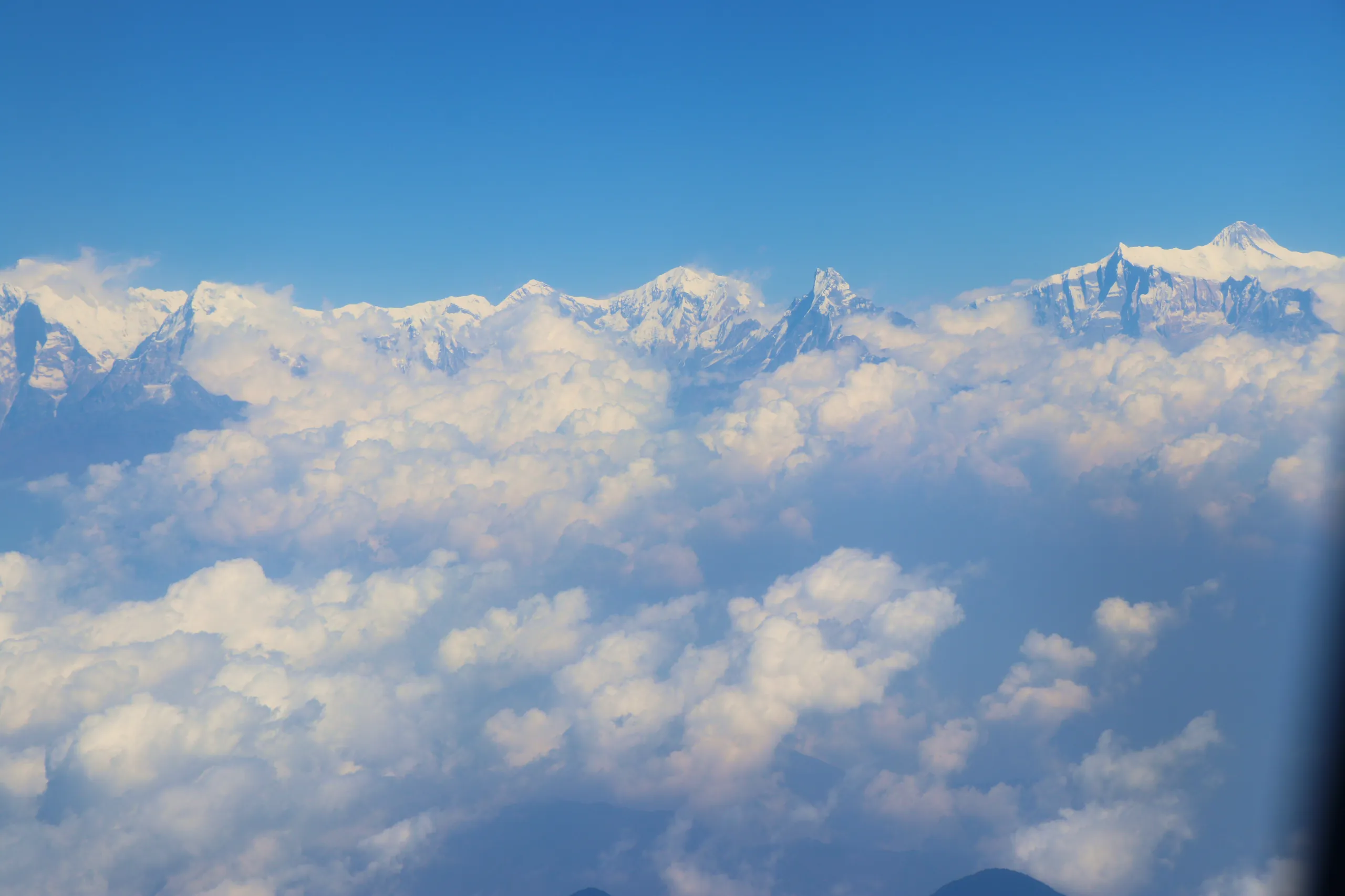 A spectacular aerial view photographed through an airplane window, looking out over the Himalayan mountain range from high altitude. The lower portion of the frame is filled with a vast, unbroken layer of white cloud that stretches to the horizon in every