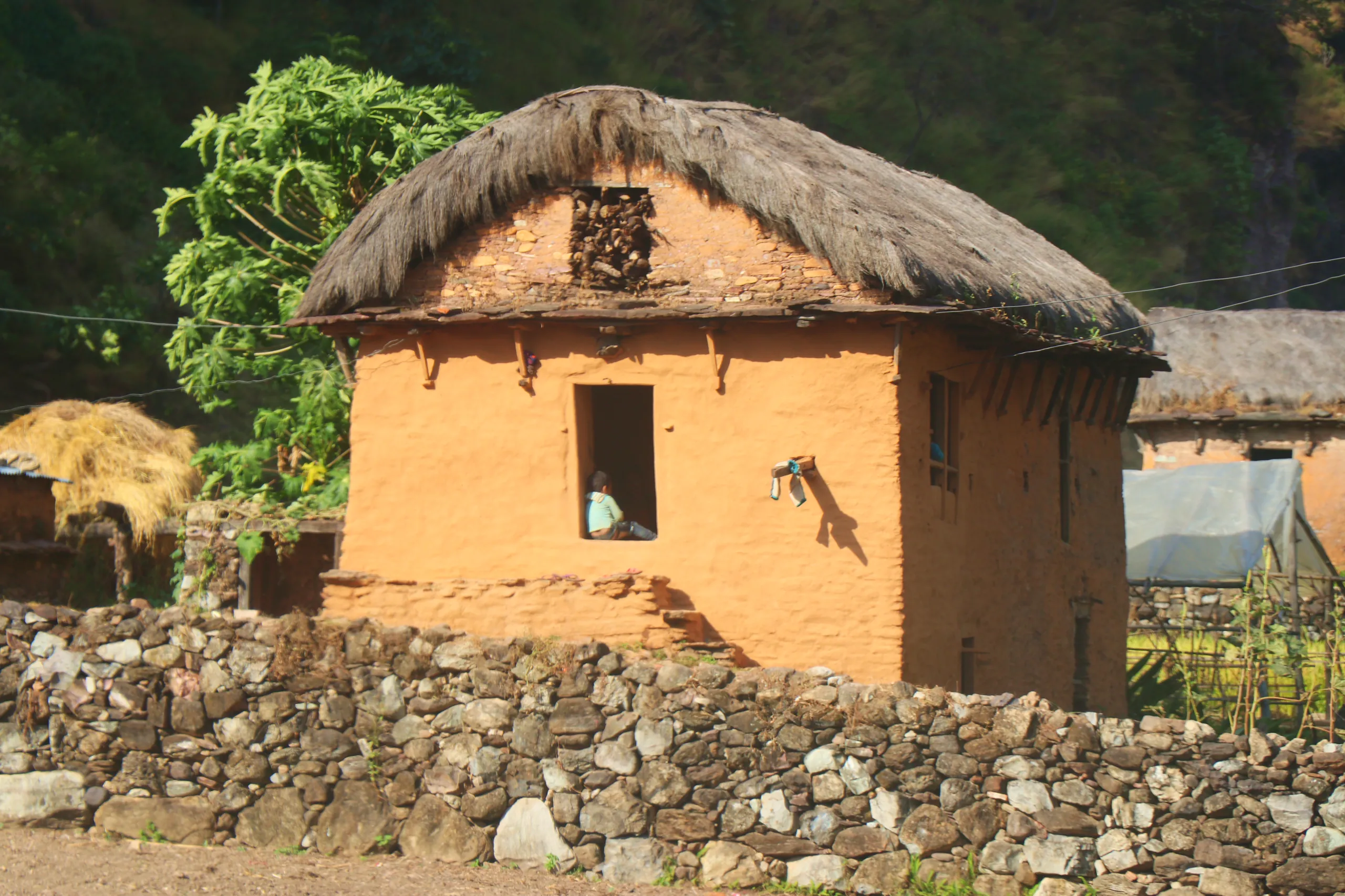 A traditional round mud-brick house with a thick, dome-like thatched roof sits in a quiet rural compound. The walls are made of smooth orange-ochre clay/mud, warm and earthy in color. A single small rectangular window opening is cut into the wall, and a p