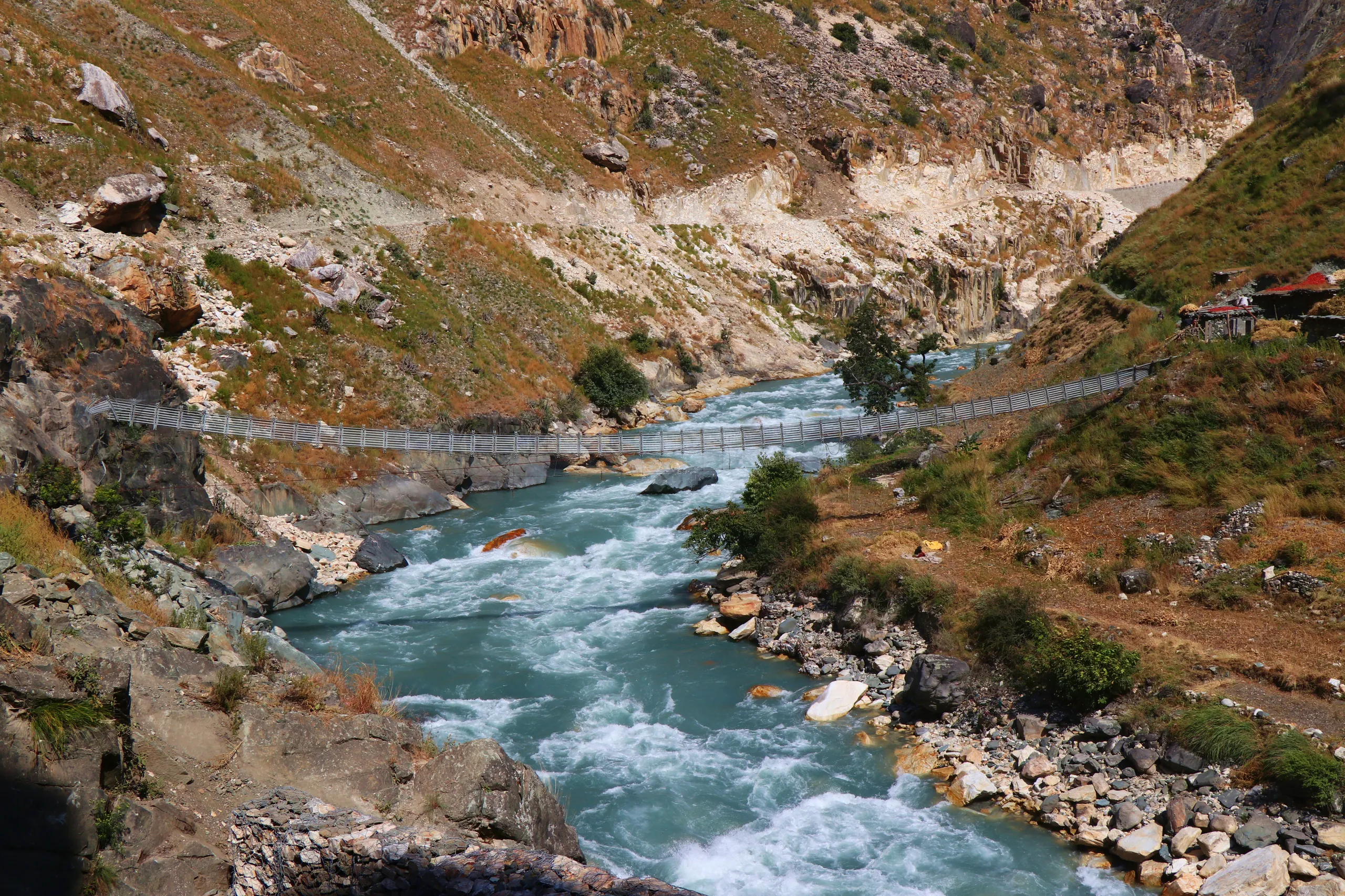 A wide mountain river gorge carved between steep, rocky, sparsely vegetated hillsides. A long suspension bridge - thin cables strung between concrete pylons - spans the wild turquoise river far below. The river rushes powerfully through the narrow gorge, 