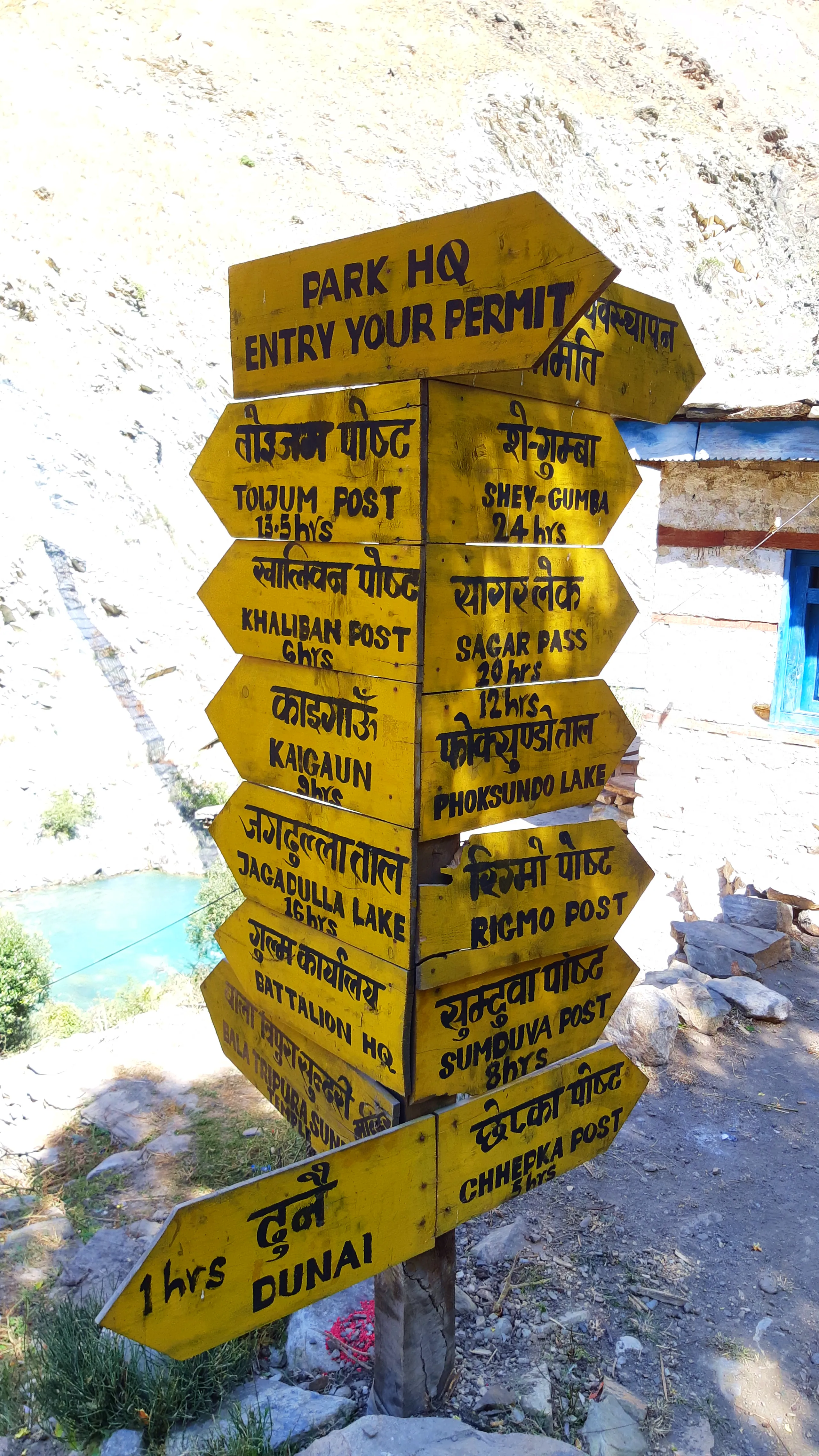 A tall wooden signpost stands in front of a rocky, sun-bleached wall, with a small blue-painted structure visible at the right edge. The post is crowded with many yellow arrow-shaped wooden signs pointing in different directions - left and right - each pa