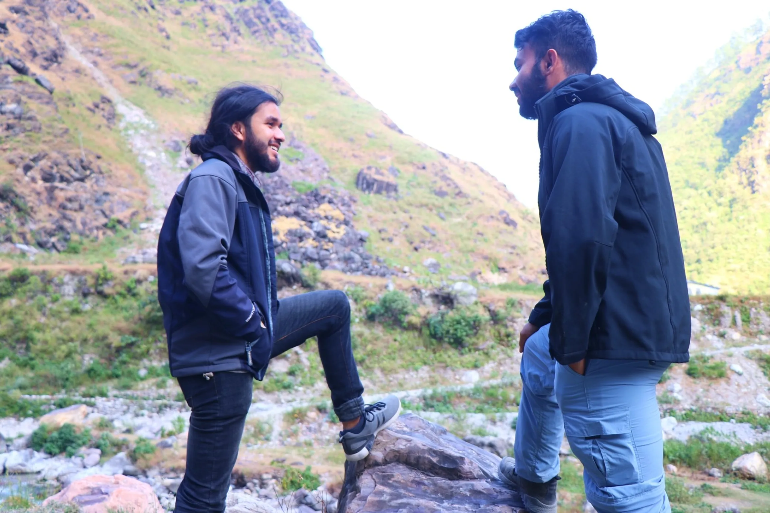 Two South Asian male trekkers engaged in an animated conversation while standing on rocks beside a wide, lush green Himalayan valley, both wearing dark trekking jackets - one in black with long dark hair tied back and a beard laughing openly while resting one foot on a boulder, the other in a plain dark hoodie with light grey cargo pants standing face-to-face, dramatic terraced green hillside and mountain ridgeline stretching across the background under a bright white sky, Nepal mountain trek camaraderie, Shey Phoksundo trekking companions, Himalayan valley conversation, friends trekking Nepal, adventure travel bonding