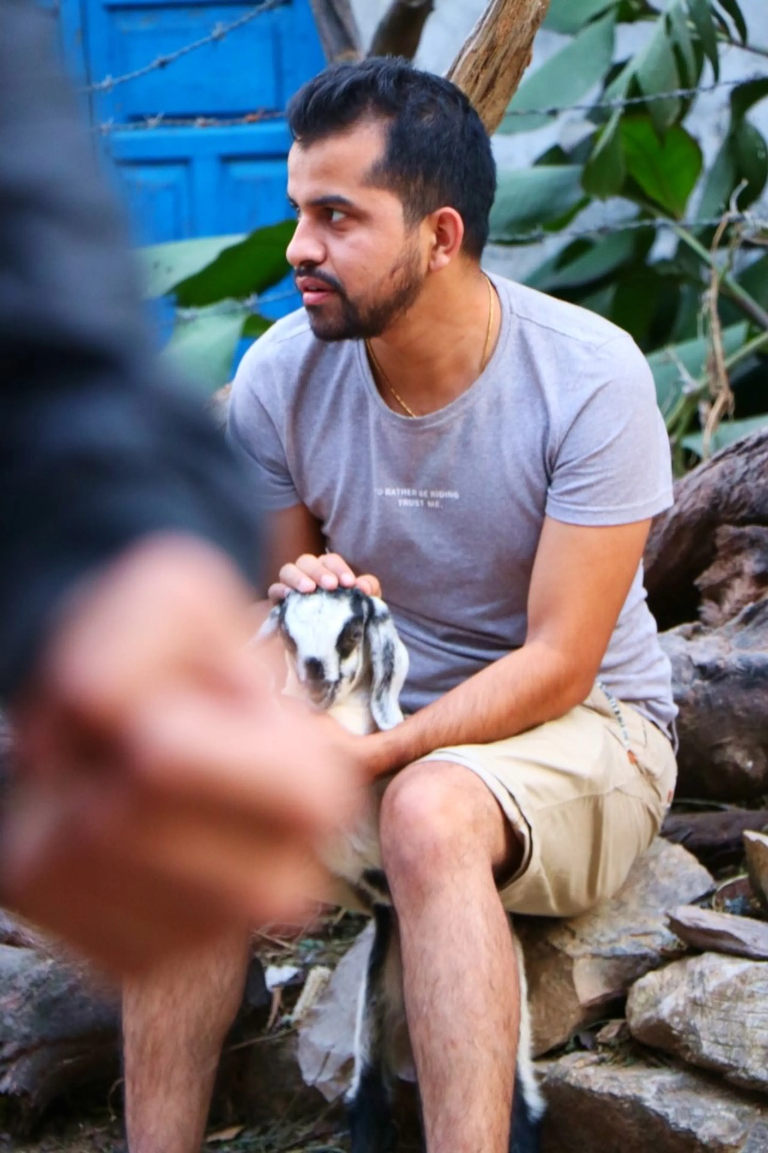 A young South Asian man with a short beard sitting on a stone wall outdoors cradling a small black and white baby goat or kid in his hands, wearing a light grey graphic t-shirt reading "I'd rather be riding, trust me" and beige khaki shorts with a thin gold chain necklace, surrounded by fallen logs and large tropical leaves against a vivid blue painted wooden door background, a blurred foreground element partially obscuring the lower left, baby goat Nepal village, trekker with animal Nepal, Shey Phoksundo trek village life, Himalayan village encounter, authentic Nepal culture trekking