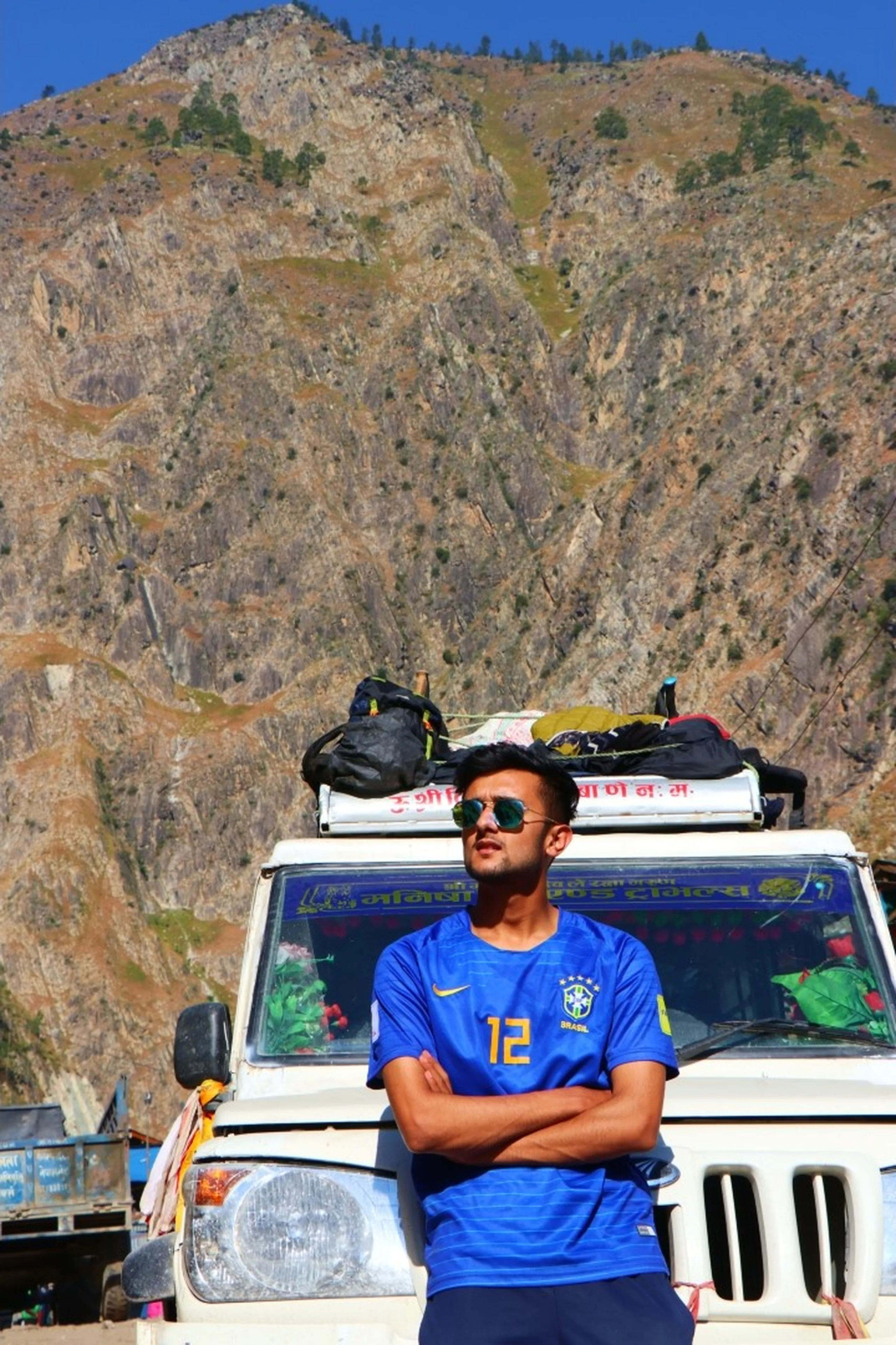 A young slim South Asian male trekker posing with arms crossed and a distant gaze in front of a white Nepali Bolero jeep loaded with gear on the roof rack, wearing a blue Brazil national football team Nike jersey with the number 12 and "BRASIL" logo, grey sports shorts and green mirrored aviator sunglasses, massive dramatic bare rocky Himalayan peak rising steeply behind in a clear blue sky, Nepali Devanagari text on jeep windshield, Nepal trekking footballer style, Brazil jersey trek Nepal mountains, Shey Phoksundo trek adventure, cool trekker Nepal Himalaya, mountain road trip Nepal