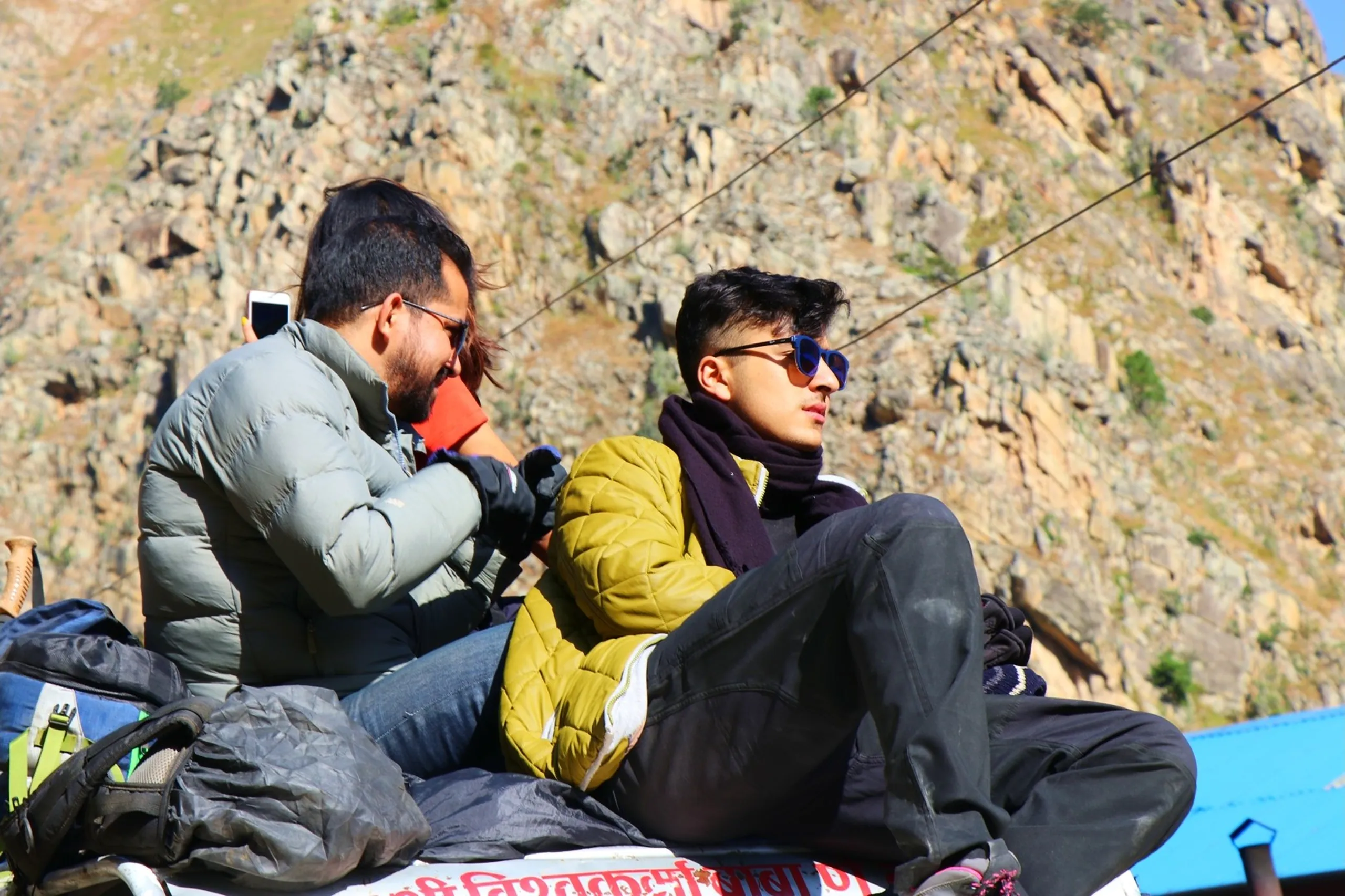 Two South Asian male trekkers sitting side by side on top of a jeep roof rack against a stark rocky Himalayan cliff backdrop under a bright sky - the one on the left with a beard wearing a silver-grey puffer jacket and dark jeans looking down at a smartphone, and the younger one on the right in an olive-yellow quilted puffer jacket with a dark scarf and blue rectangular sunglasses gazing stoically into the distance, overhead power lines crossing the frame, blue building corner visible below, Nepali text on jeep, rooftop jeep ride Nepal Himalaya, Shey Phoksundo trek journey, trekking companions Nepal mountain road, Himalayan adventure travel