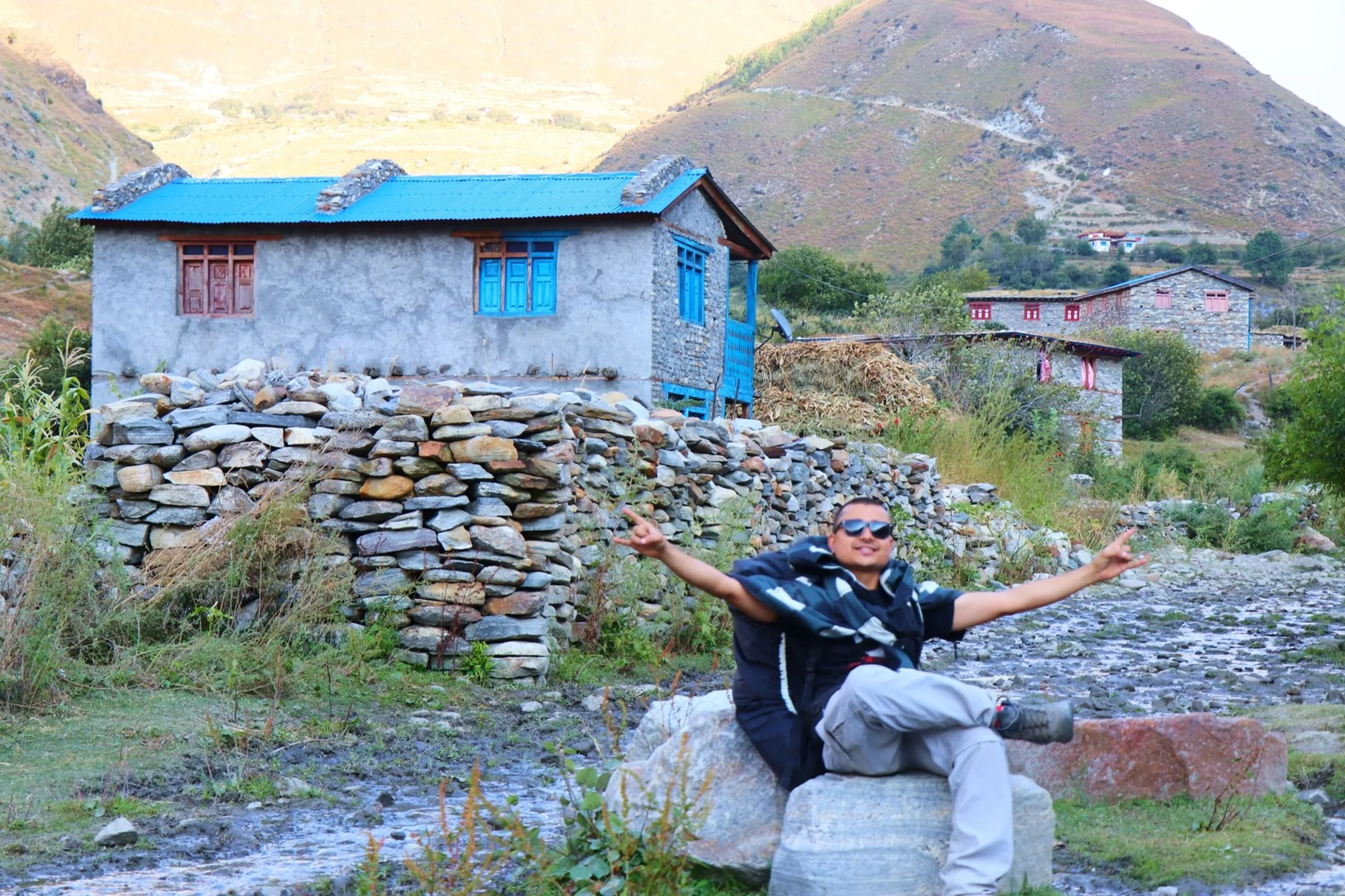 A young South Asian male trekker lounging joyfully on a large flat boulder with arms spread wide and fingers making peace or rock-on signs, wearing dark sunglasses, a black jacket and light grey trekking trousers with hiking boots, a joyful and carefree expression on his face, set against a picturesque traditional Nepali mountain village scene - stone-walled enclosures, a grey stone house with bright blue corrugated tin roof and matching blue window frames, golden hillside and rocky peaks in the background, a small stream or puddle visible on the ground, Nepal mountain village photography, Shey Phoksundo remote village trek, traditional Nepali stone house, Himalayan trekking joy, authentic Nepal travel photography