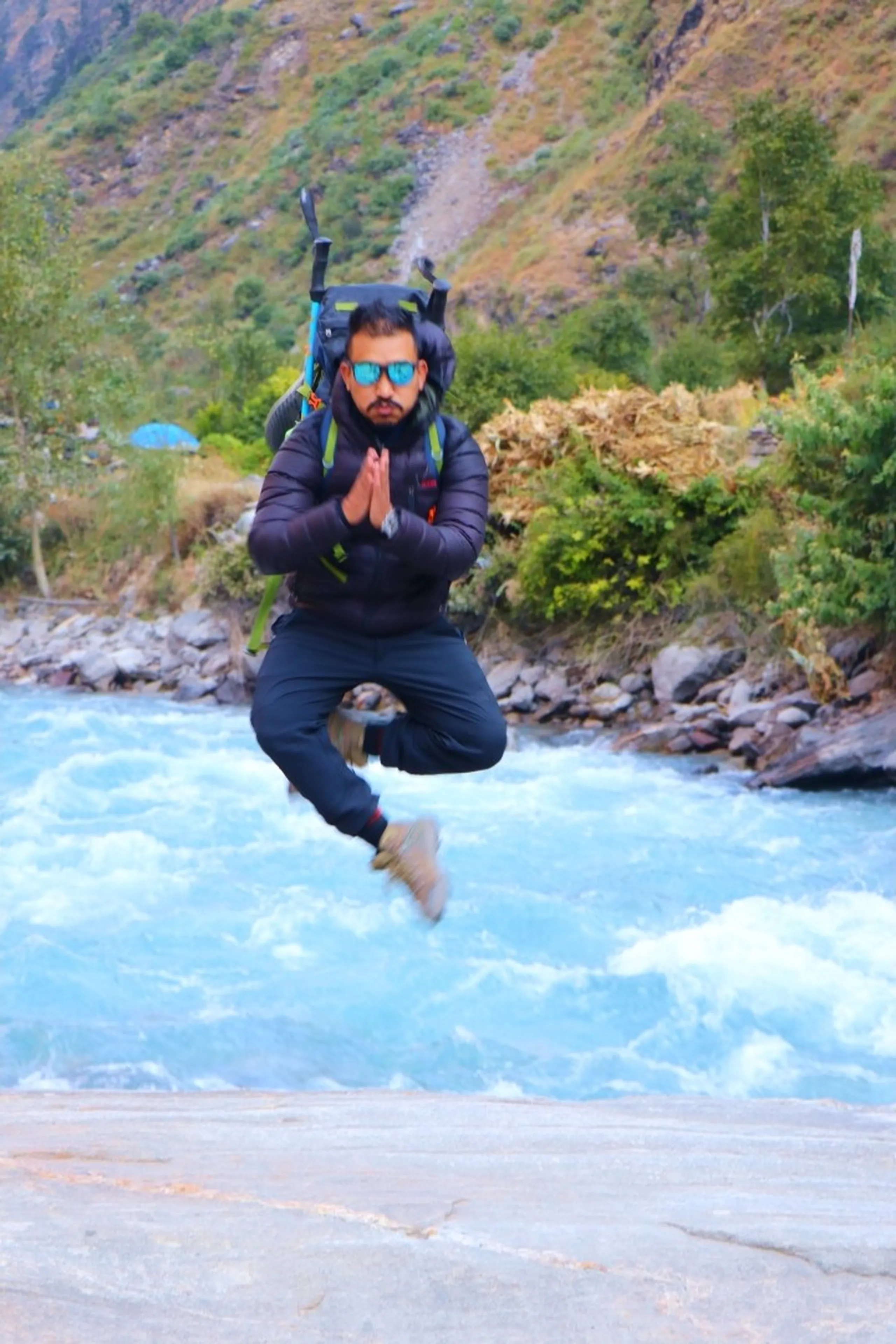 A male trekker captured mid-air in a dramatic levitation jump shot over a stone bridge with a raging turquoise glacial river in the background, wearing a black puffer down jacket, black trekking pants and tan/beige hiking boots, a large green-accented hiking backpack with trekking poles sticking up, hands pressed together in a Namaste prayer gesture with a serious expression and teal mirrored sunglasses, green forested hillside behind, blue prayer flags barely visible in the distance, Nepal jump shot photography, Namaste jump river Nepal, Shey Phoksundo suspension bridge trekking, glacial river Nepal Himalaya, viral trekking photo Nepal, adventure photography Nepal mountains